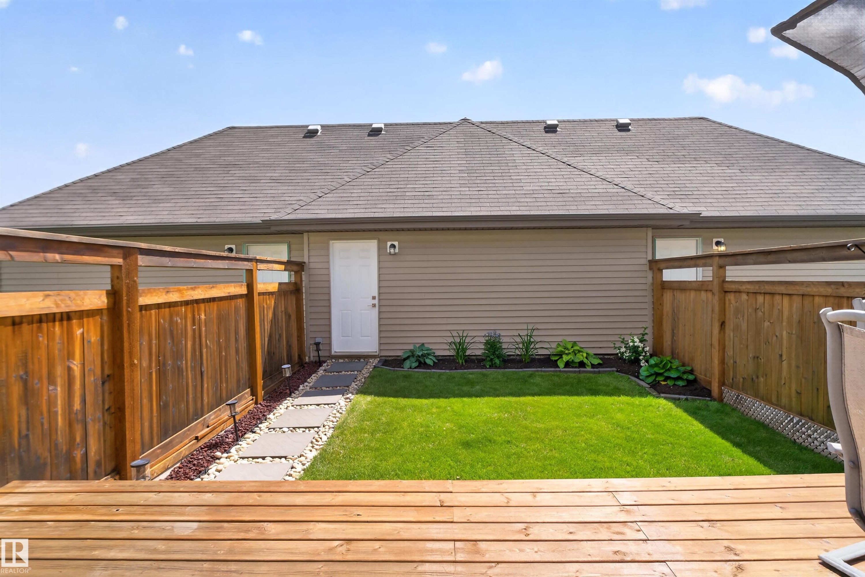Expansive wooden deck leading to a fenced backyard with a manicured lawn, stone pathway, and landscaped garden beds - 319 Desrochers Boulevard Sw, Edmonton, AB