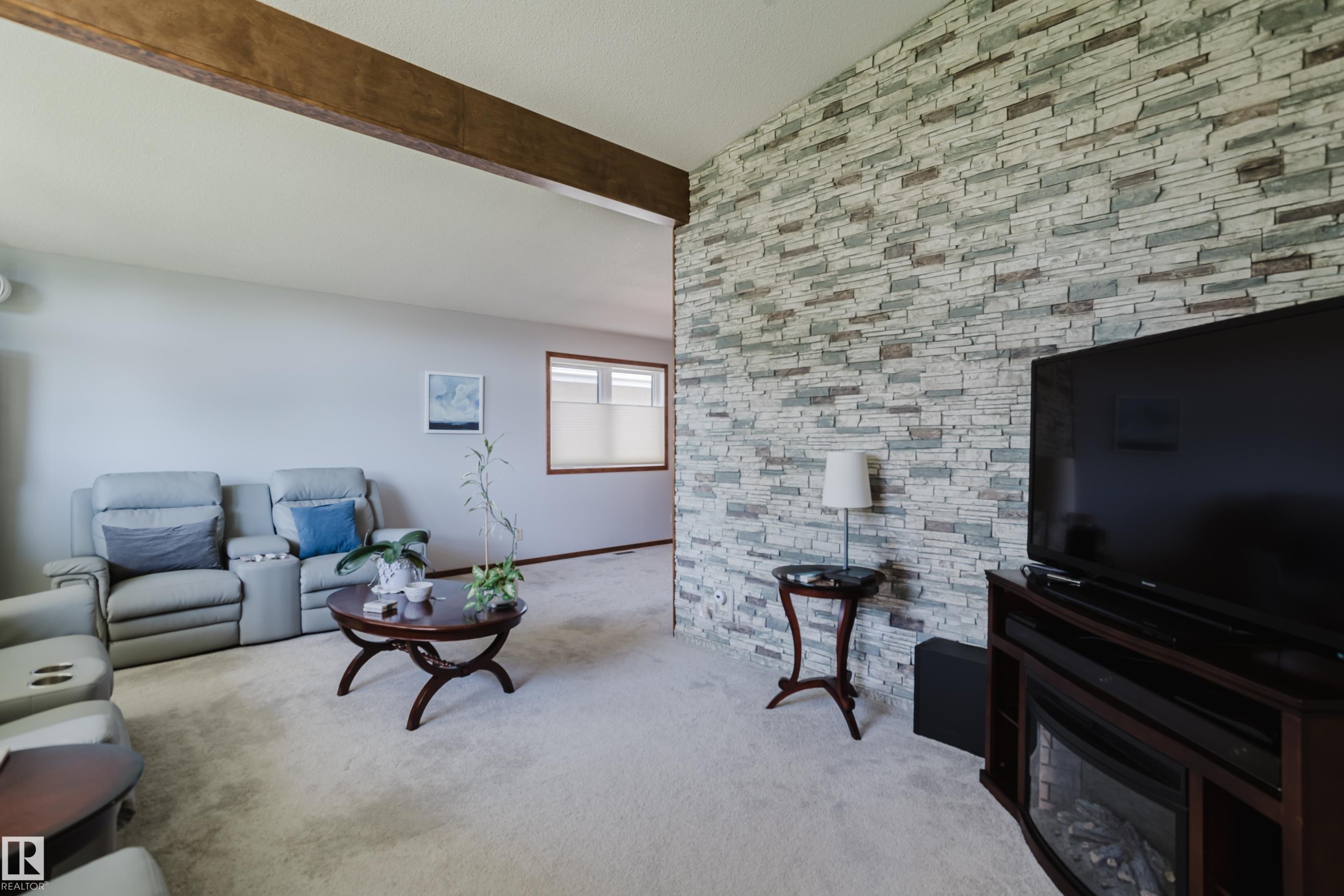 Exposed wood beam, light-toned wall-to-wall carpeting, window with a privacy blind, and a stacked stone accent wall with an integrated electric fireplace - 10924 35A Avenue Nw, Edmonton, AB