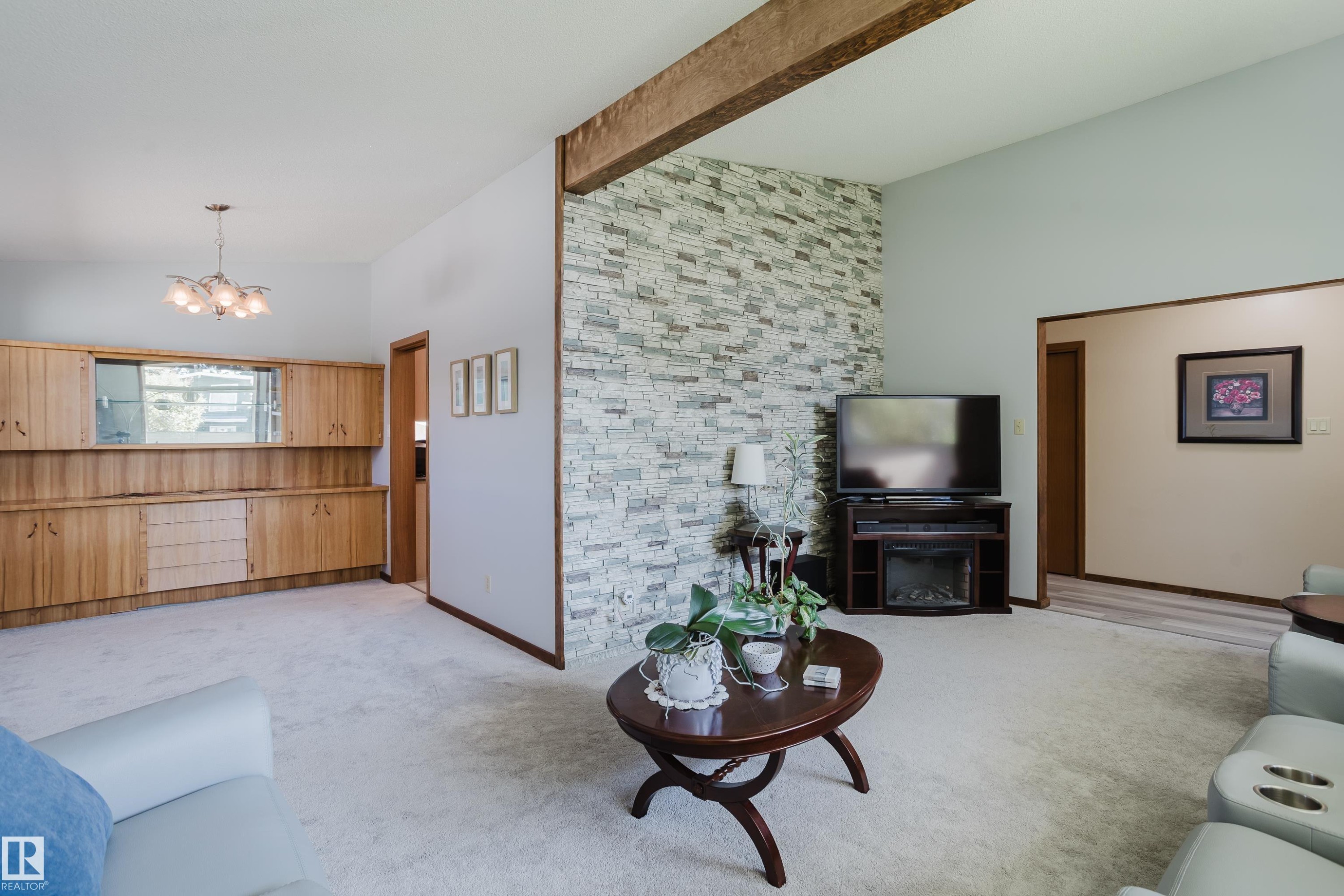 Vaulted ceiling with exposed wood beam, built-in natural wood cabinetry, stone accent wall, recessed electric fireplace, and a contemporary chandelier - 10924 35A Avenue Nw, Edmonton, AB