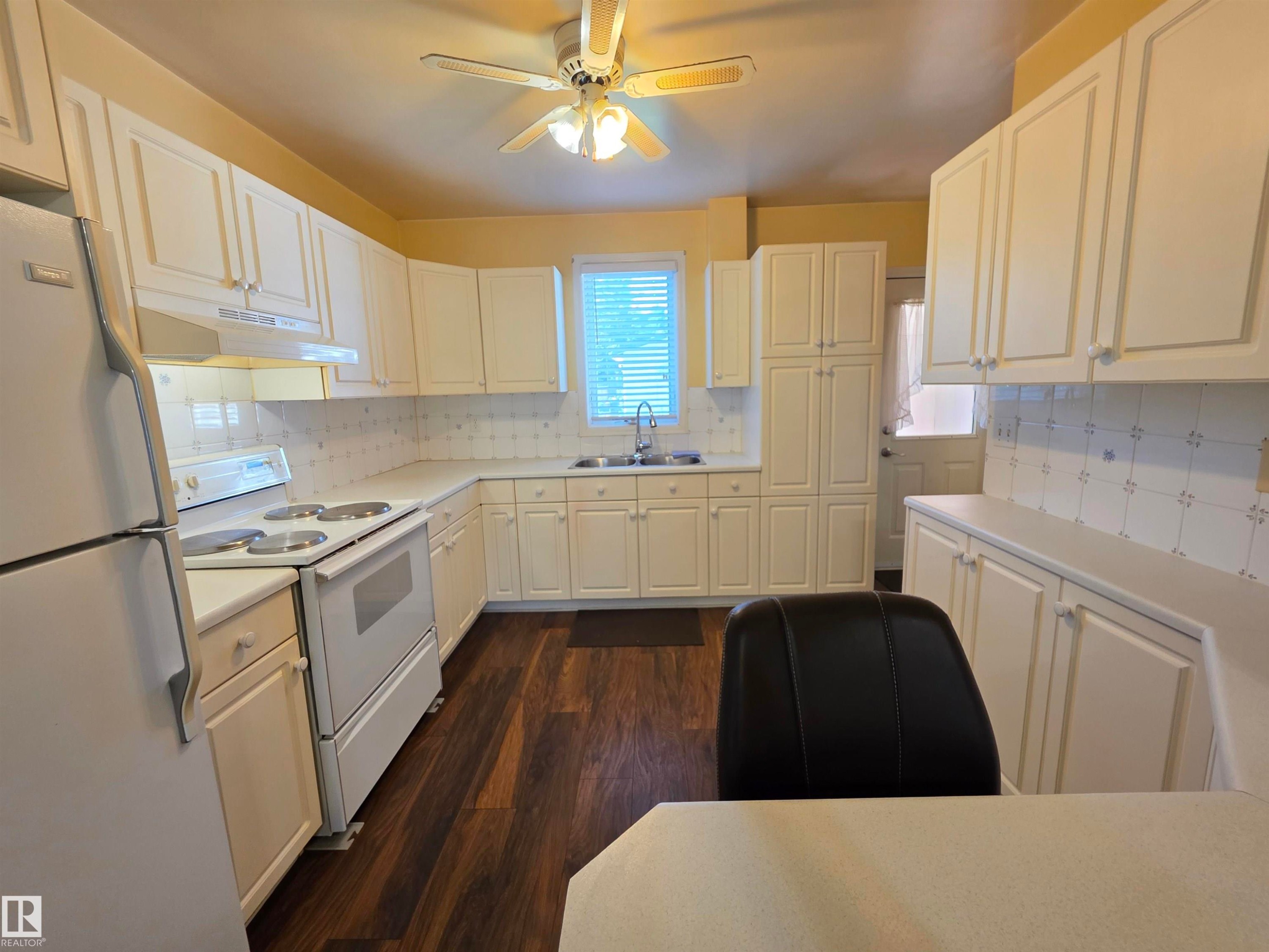 Kitchen featuring extensive white cabinetry, wood-finish flooring, a ceiling fan with integrated lighting, and a white tile backsplash - 10438 147 Street Nw, Edmonton, AB