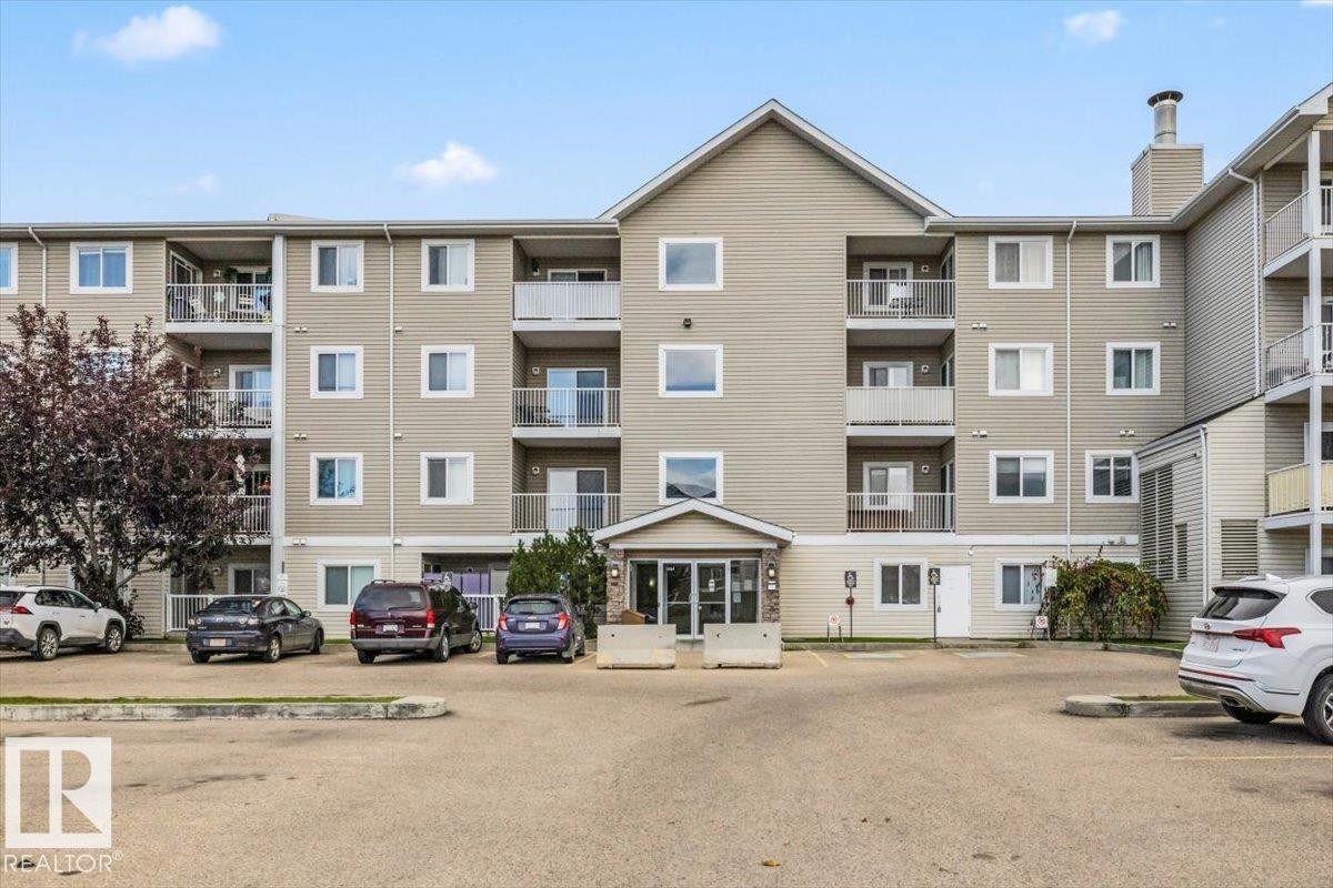 Multi-story building with neutral-toned siding, multiple balconies with glass railings, and a prominent peaked roofline - #110 4309 33 St, Stony Plain, AB