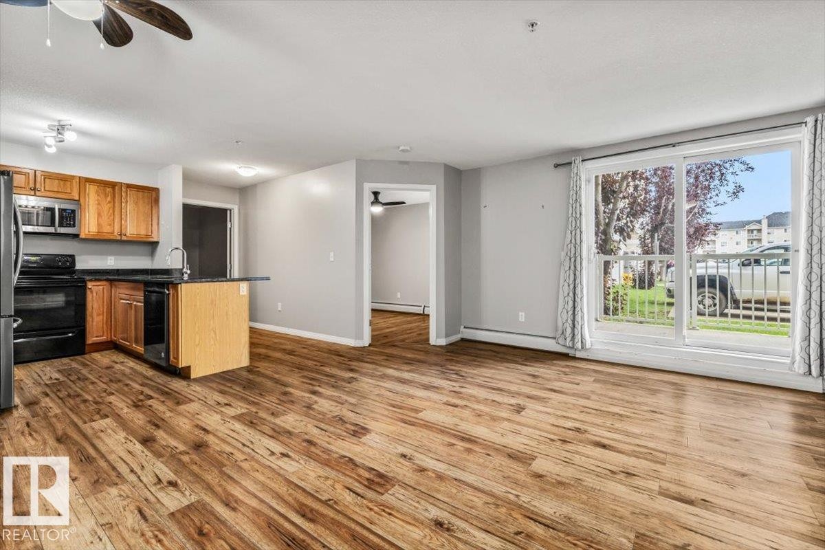 Expansive main living area featuring wood-finish flooring, a kitchen with wood cabinetry and black appliances, an island with a sink, and a large sliding glass door - #110 4309 33 St, Stony Plain, AB
