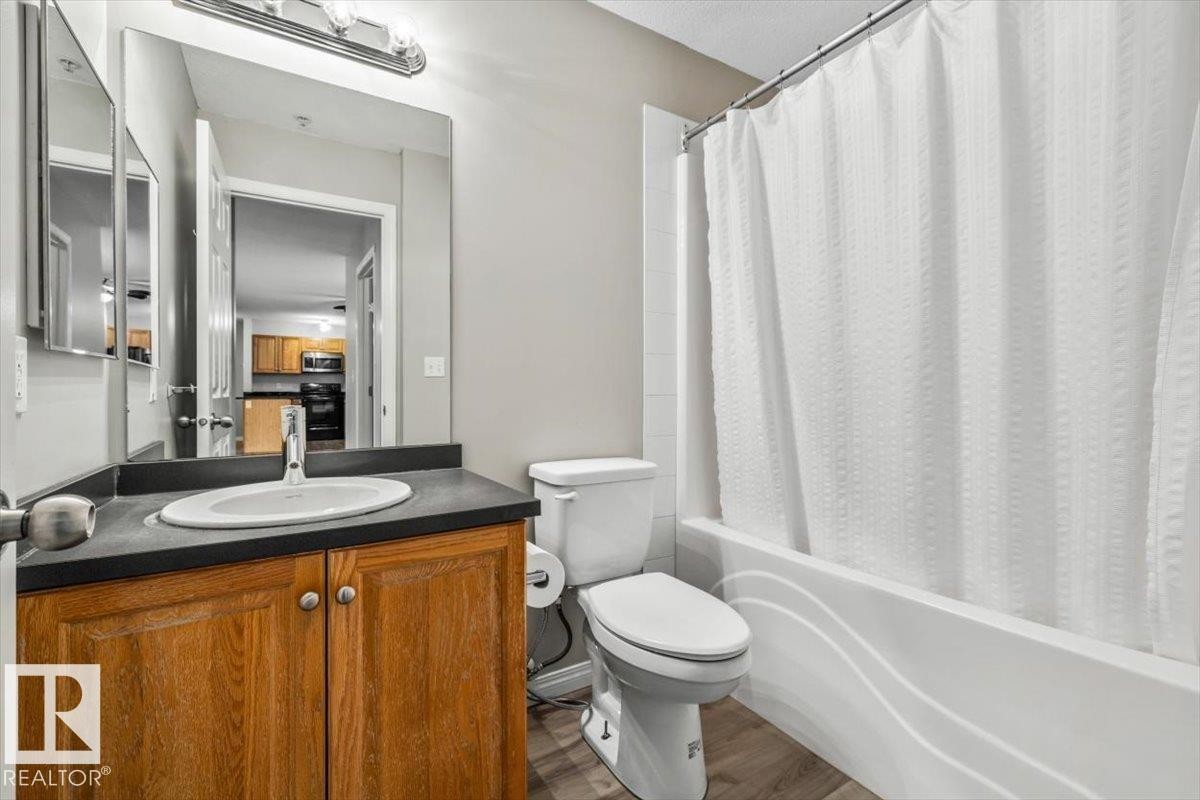 Bathroom featuring a wood-finish vanity with an integrated sink, a dark countertop, an expansive mirror, and a white bathtub with tile surround - #110 4309 33 St, Stony Plain, AB
