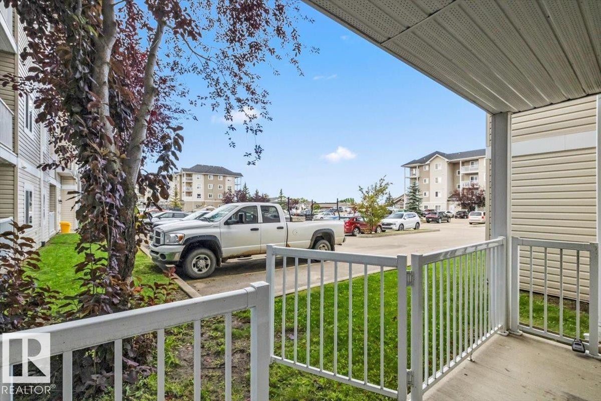 Covered patio with white railing, concrete floor, and adjacent green space - #110 4309 33 St, Stony Plain, AB