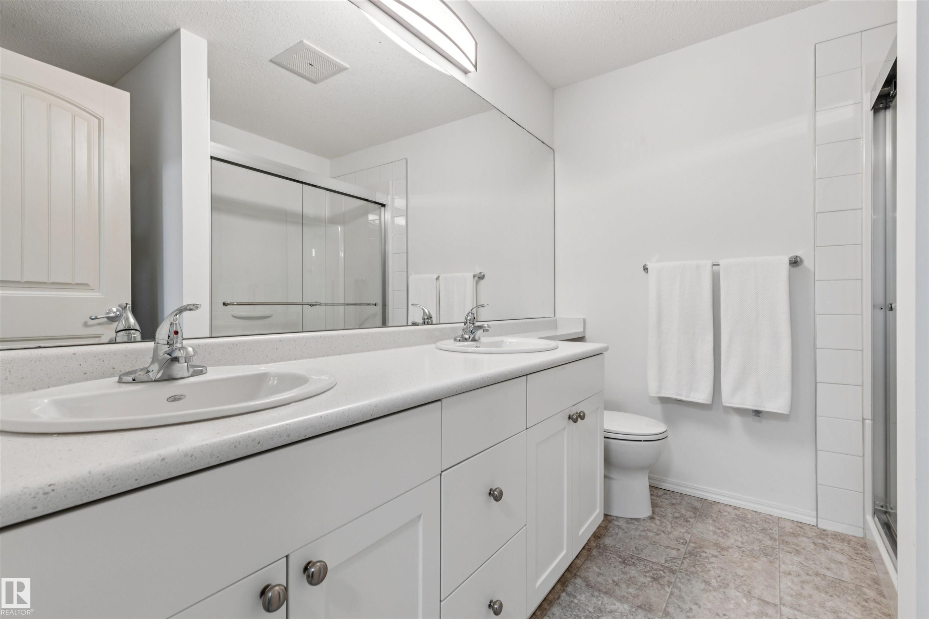 Dual vanity with integrated sinks and chrome faucets, full-width mirror, white cabinetry, and adjacent toilet - #353 2096 Blackmud Creek Dr Sw, Edmonton, AB - Indoor Photo Showing Bathroom