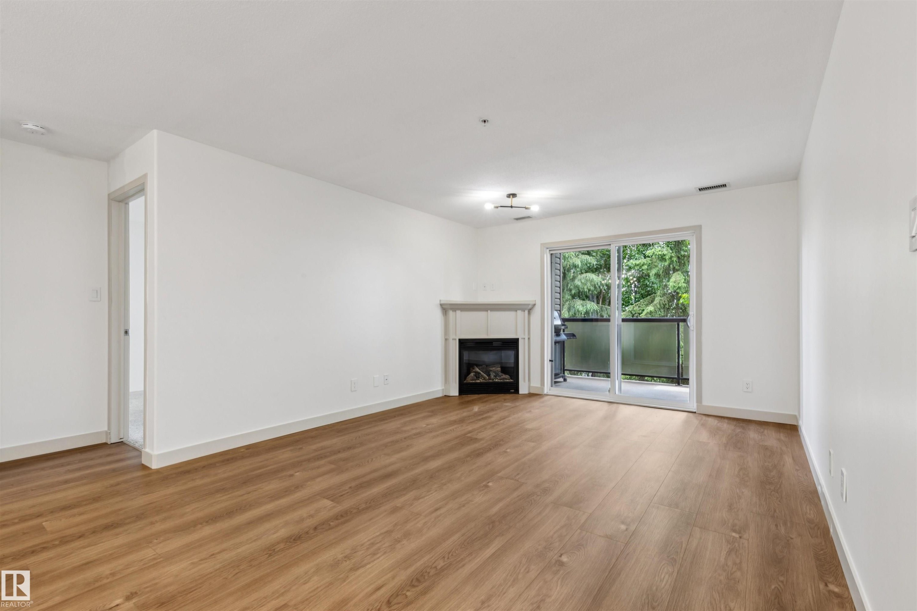 Spacious living area featuring wood-finish flooring, a contemporary fireplace with mantel, and a sliding glass door opening to an outdoor balcony with a glass railing - #353 2096 Blackmud Creek Dr Sw, Edmonton, AB - Indoor Photo Showing Living Room With Fireplace
