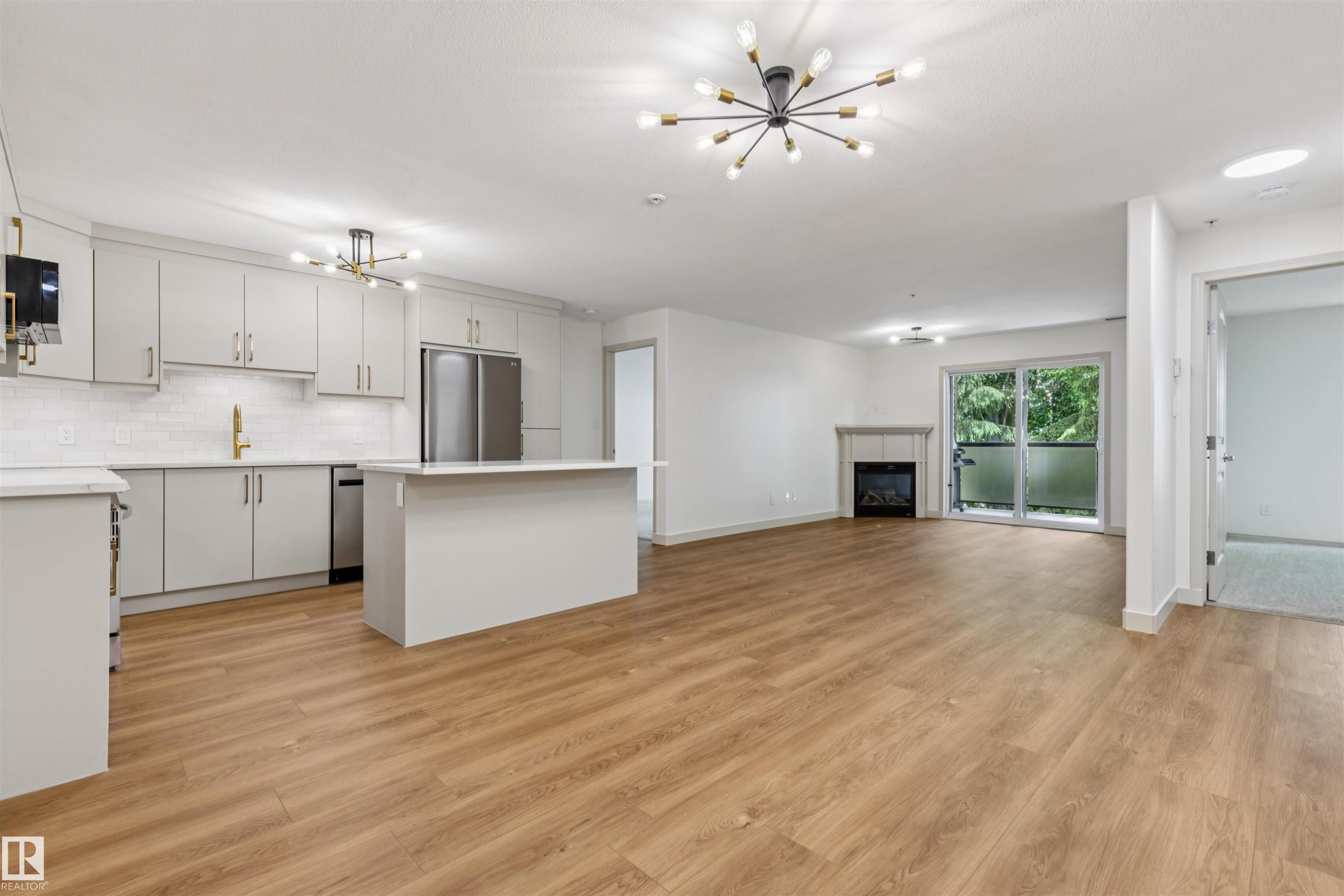Open-concept living space featuring wood-finish flooring, a contemporary kitchen with light-colored cabinetry and an island, stainless steel appliances, a tile backsplash, and a fireplace with a mantel - #353 2096 Blackmud Creek Dr Sw, Edmonton, AB - Indoor Photo Showing Kitchen With Upgraded Kitchen