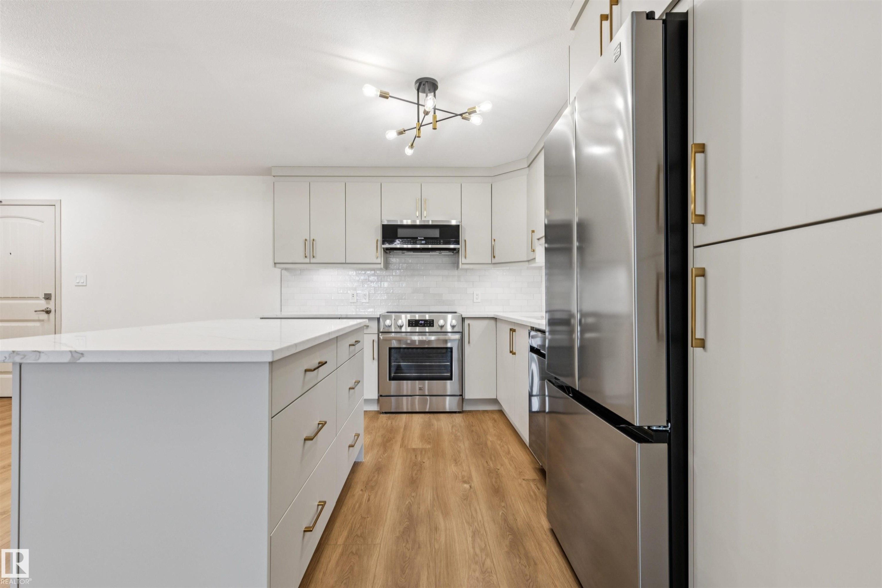 Kitchen featuring a spacious island with a light-colored countertop, stainless steel appliances, light cabinetry with gold-toned hardware, wood-finish flooring, and a contemporary ceiling light fixture - #353 2096 Blackmud Creek Dr Sw, Edmonton, AB - Indoor Photo Showing Kitchen With Stainless Steel Kitchen With Upgraded Kitchen