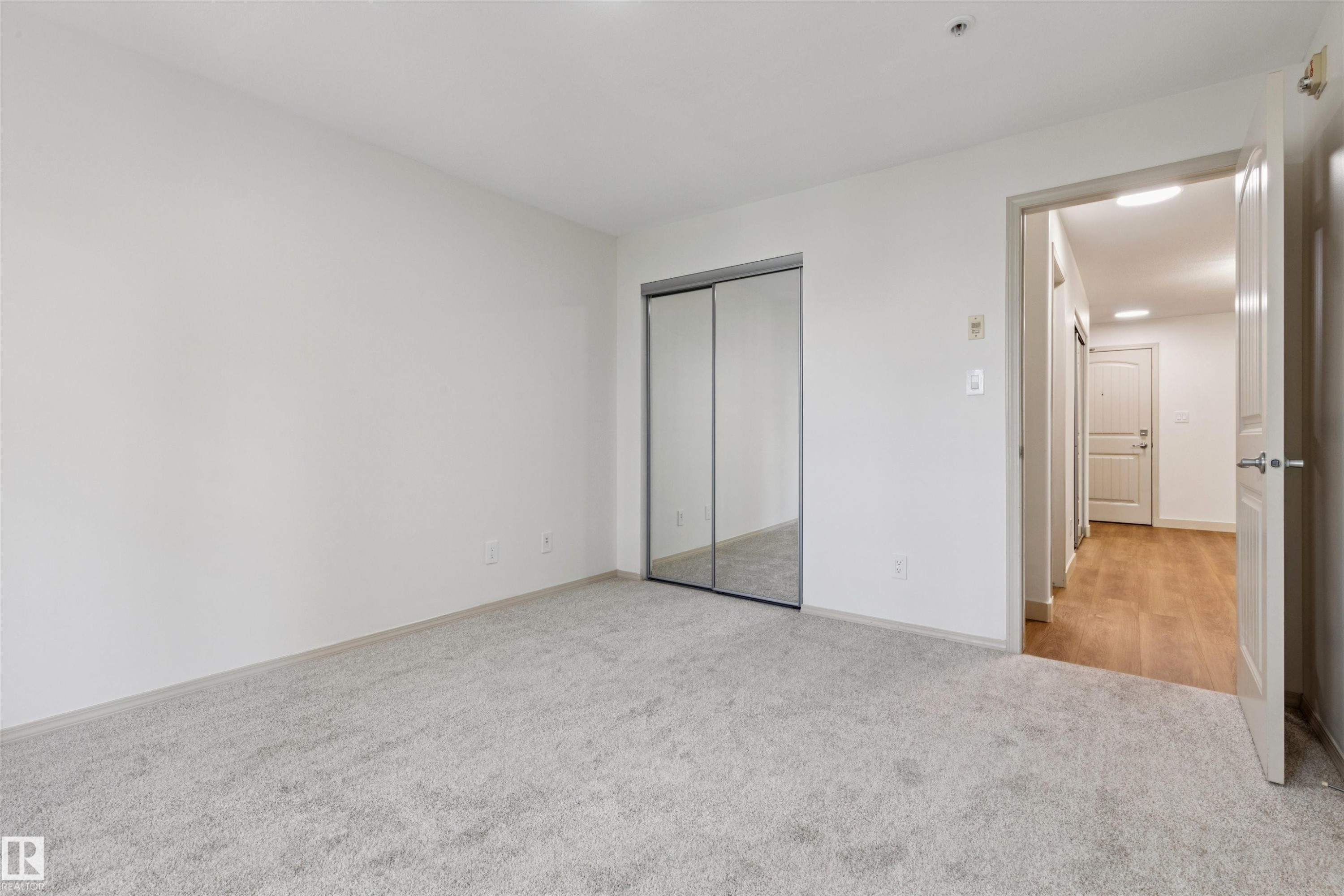 Carpeted room featuring a mirrored sliding closet door, white walls, and a doorway opening to a hallway with wood-finish flooring - #353 2096 Blackmud Creek Dr Sw, Edmonton, AB - Indoor Photo Showing Other Room