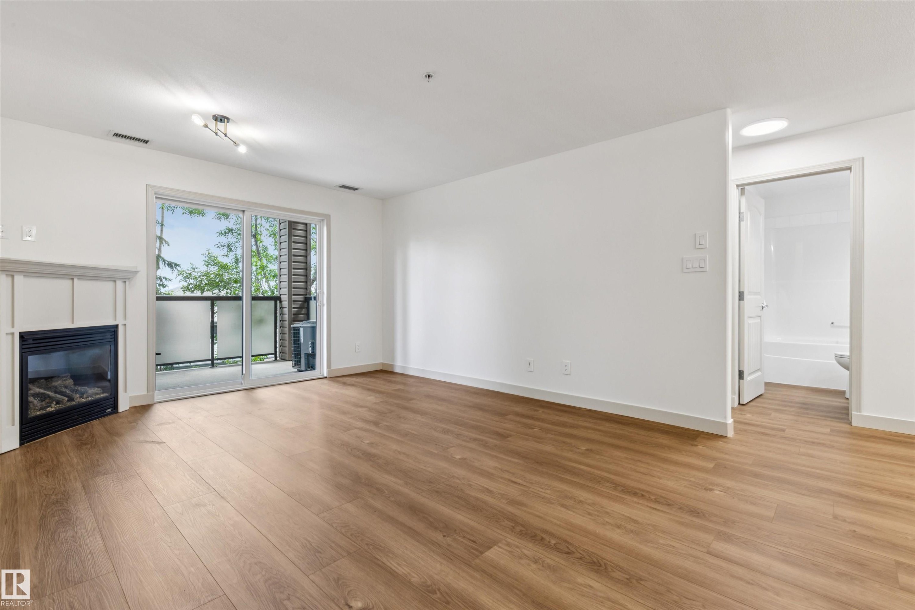 Open-concept living area featuring wood-finish flooring, a contemporary fireplace with a white mantel, and a sliding glass door opening to a private balcony - #353 2096 Blackmud Creek Dr Sw, Edmonton, AB - Indoor Photo Showing Living Room With Fireplace