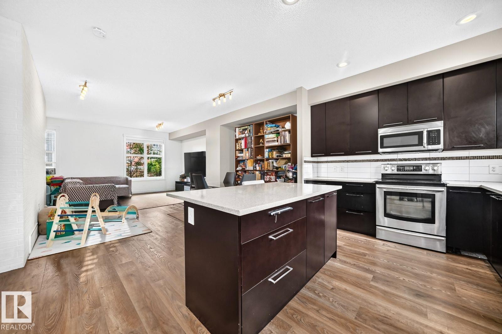 Kitchen island with a light-colored countertop, stainless steel appliances, dark wood-finish cabinetry, and wood-finish flooring - #20 8315 180 Av Nw, Edmonton, AB