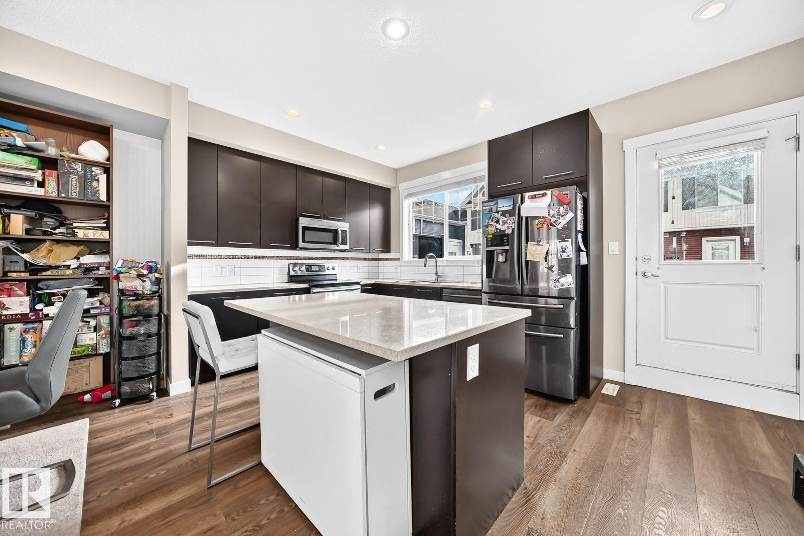Kitchen featuring wood-finish flooring, dark wood cabinetry, stainless steel appliances, a white tile backsplash, and an island with a light-colored countertop - #20 8315 180 Av Nw, Edmonton, AB