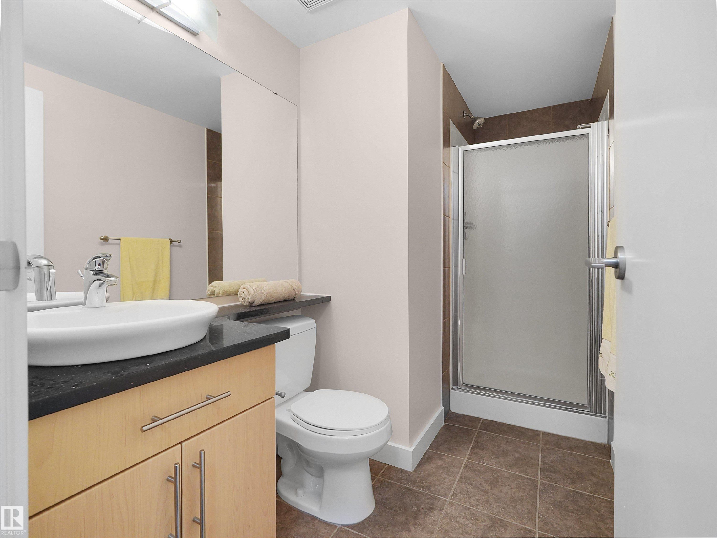 Modern vanity with a vessel sink, dark countertop, light wood-finish cabinetry, and a shower stall with a framed frosted glass door - #1601 10136 104 St Nw, Edmonton, AB