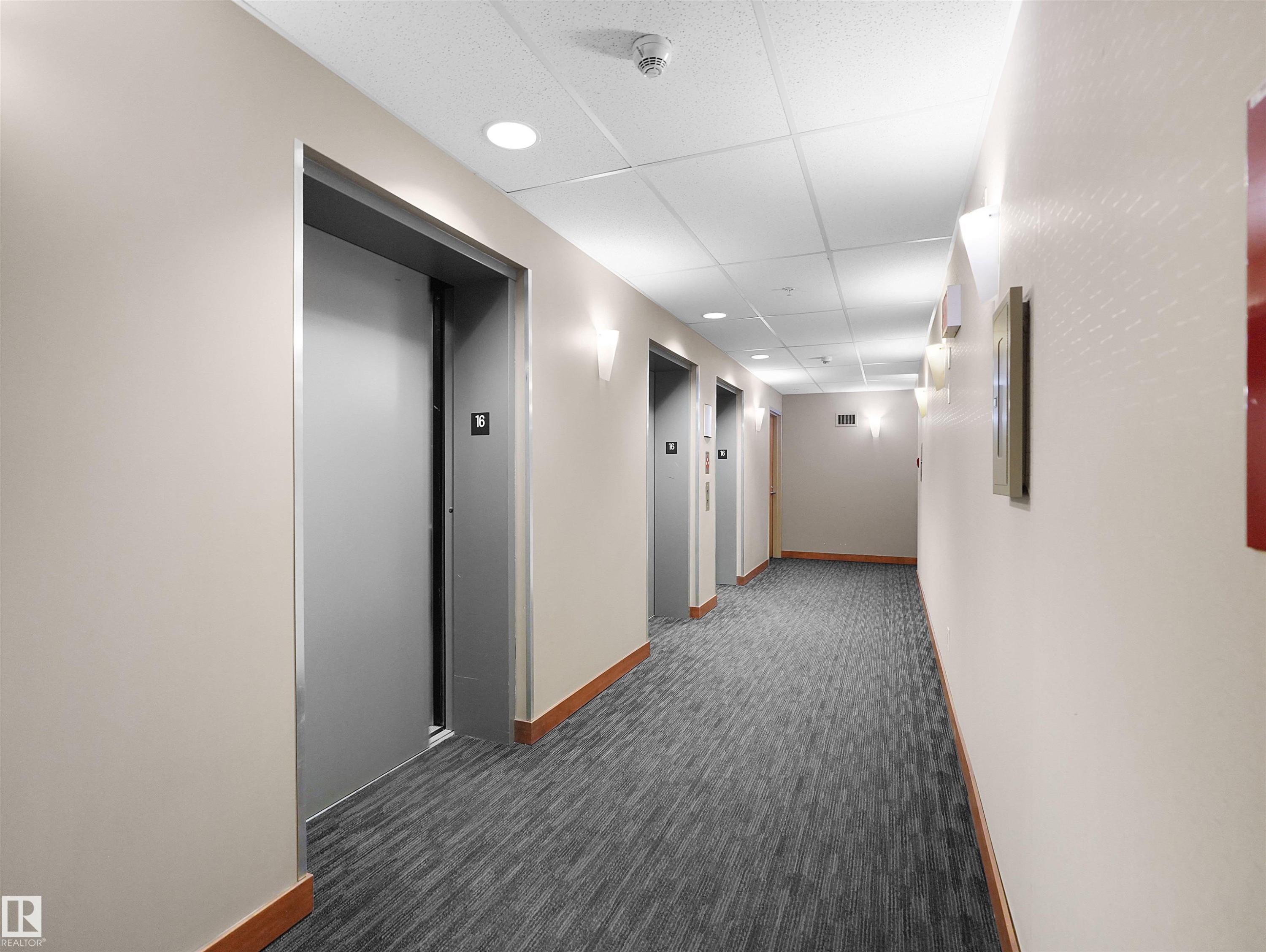 Corridor featuring multiple elevators with metallic doors, recessed ceiling lighting, wall-mounted sconces, and wood-finish baseboards - #1601 10136 104 St Nw, Edmonton, AB
