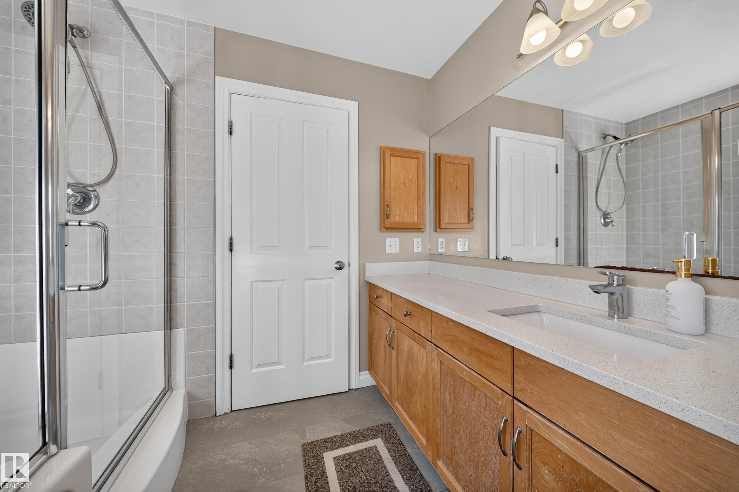 Built-in tub with glass enclosure, single vanity with light wood-finish cabinetry, integrated sink, and white countertop - 121 64 Street Sw, Edmonton, AB