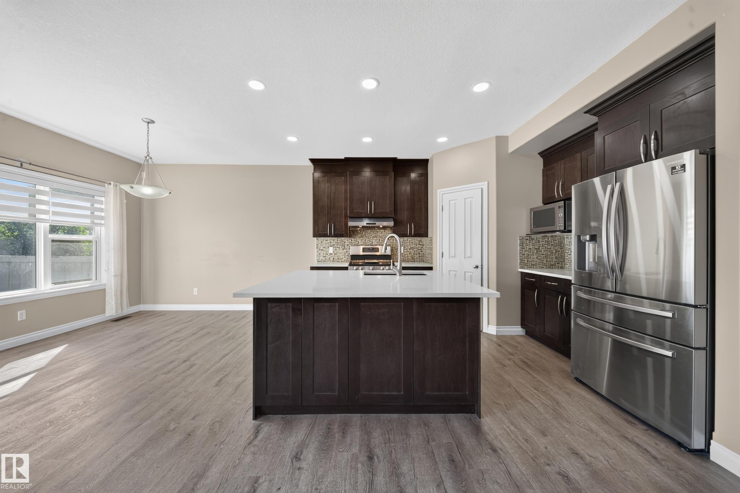 Open-concept kitchen featuring dark wood cabinetry, stainless steel appliances, a center island with a light-colored countertop, and recessed lighting - 121 64 Street Sw, Edmonton, AB