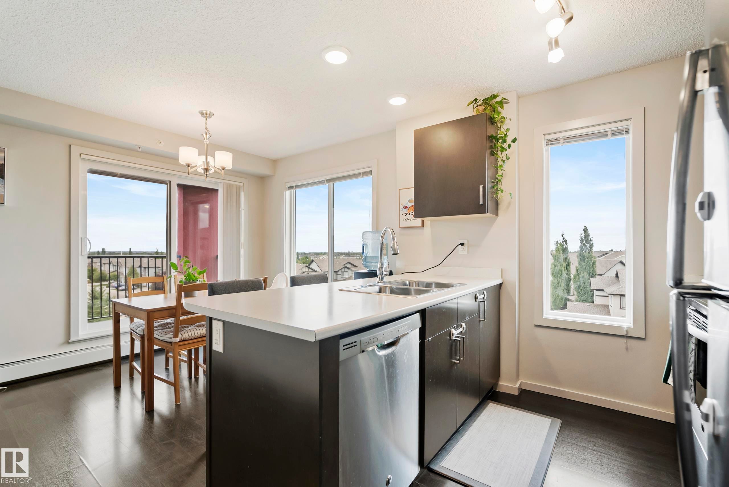 Kitchen featuring wood-finish flooring, stainless steel appliances, dark cabinetry, and an eat-in island with a white countertop - #419 5515 7 Av Sw, Edmonton, AB