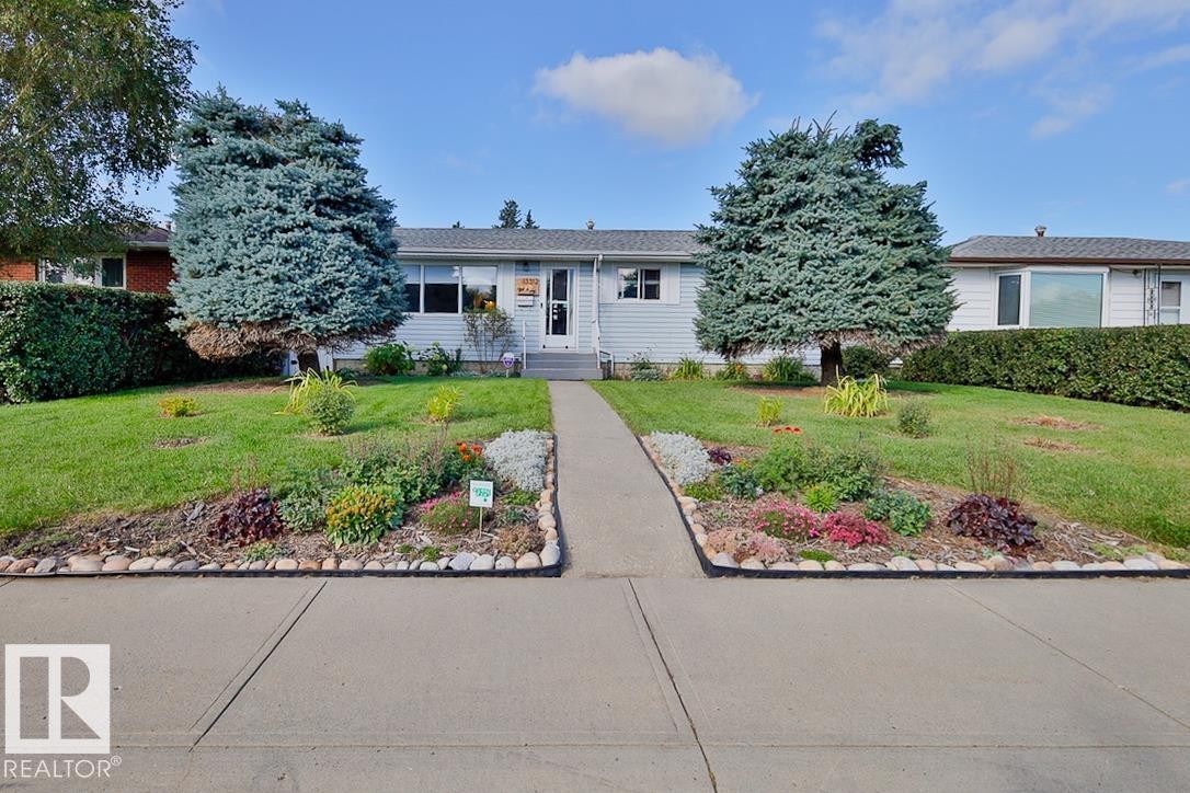 Front exterior with light blue siding, a central pathway, two mature evergreen trees, and landscaped garden beds - 13312 123A Street Nw, Edmonton, AB