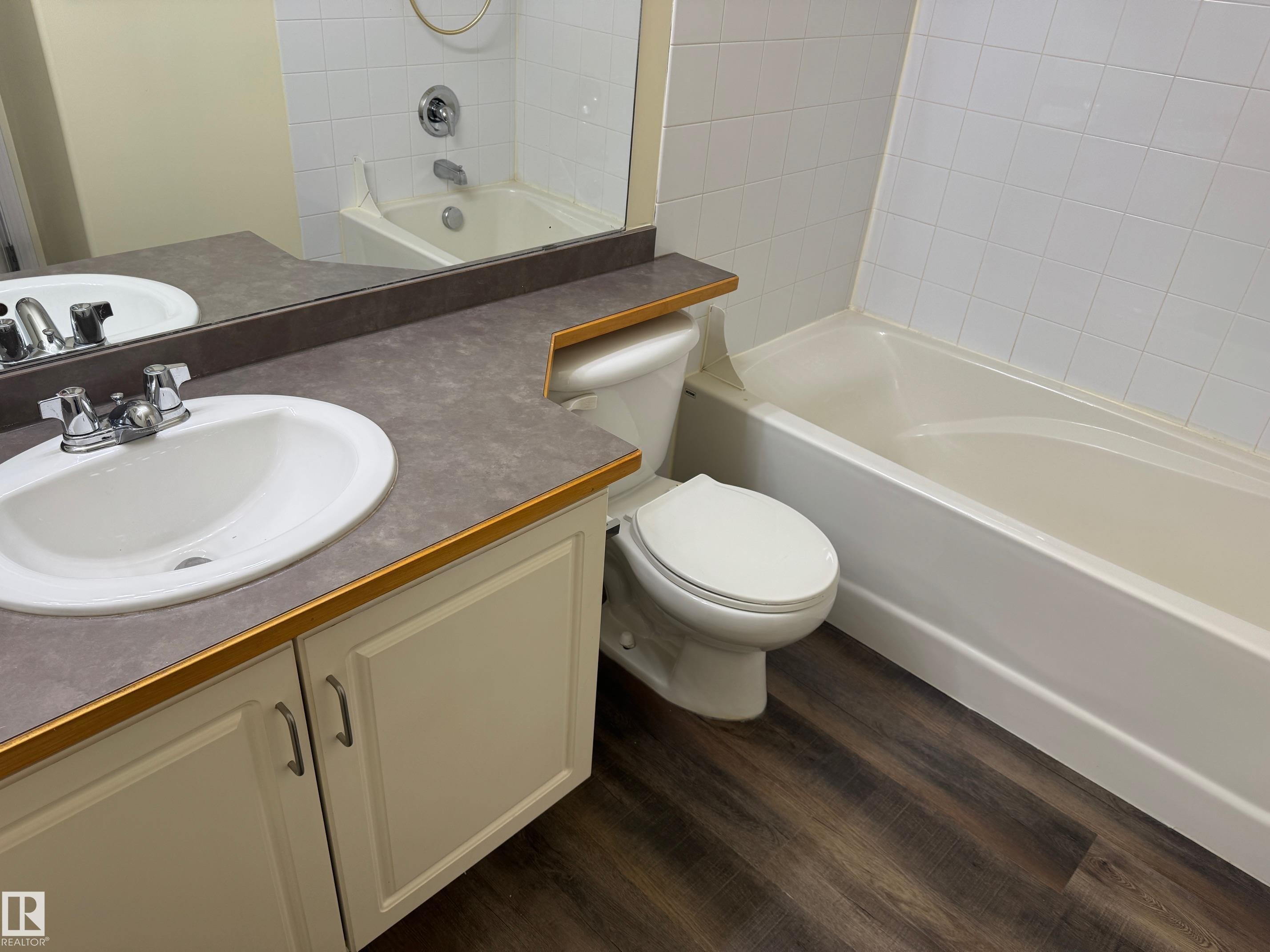 Bathroom featuring a built-in vanity with an integrated sink, a full-width mirror, a toilet, a bathtub with a tiled surround, and wood-finish flooring - #229 2305 35A Av Nw, Edmonton, AB