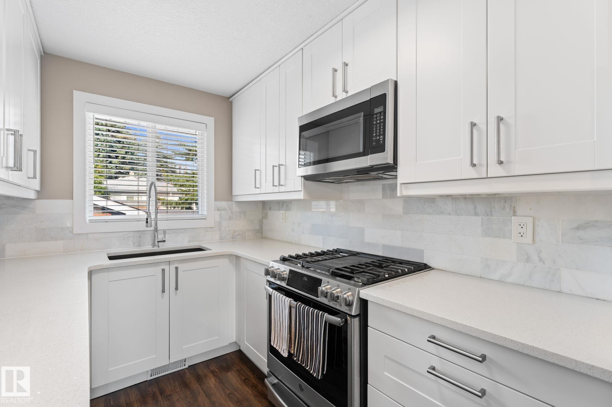 Kitchen featuring white shaker-style cabinetry, stainless steel appliances, light-toned quartz countertops, and a full-height backsplash - 6108 102 Avenue Nw, Edmonton, AB