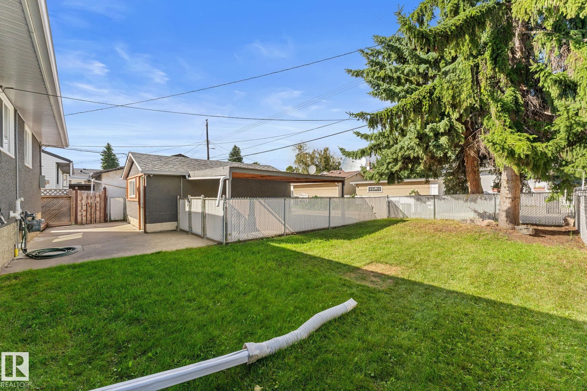 Expansive backyard with a green lawn, concrete patio, a detached detached single garage, single covered carport, chain-link fencing, and mature trees - 6108 102 Avenue Nw, Edmonton, AB