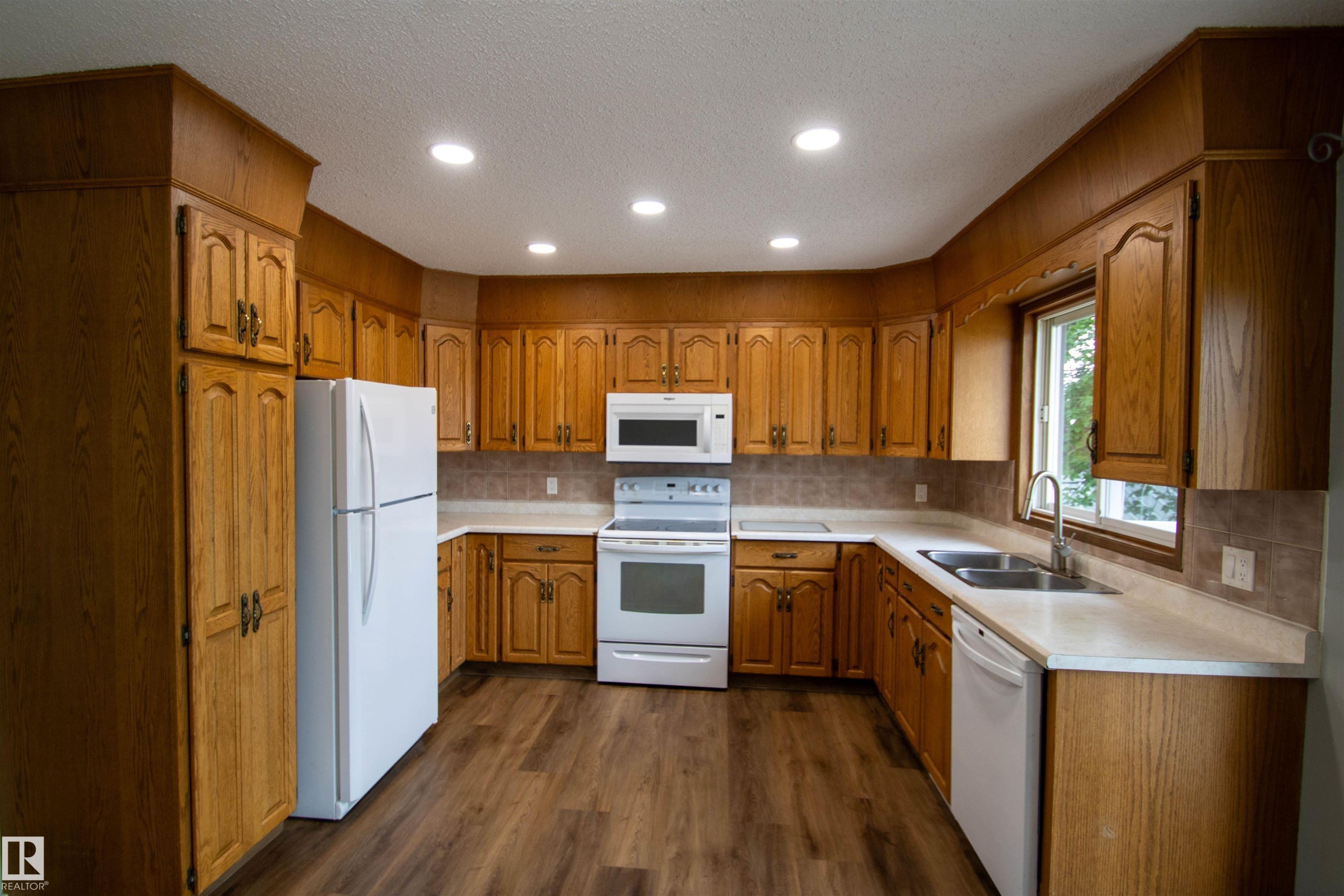 Kitchen featuring wood cabinetry, white appliances, recessed lighting, wood-finish flooring, and a window above the sink - 4725 45 Ave, St. Paul Town, AB - Indoor Photo Showing Kitchen With Double Sink