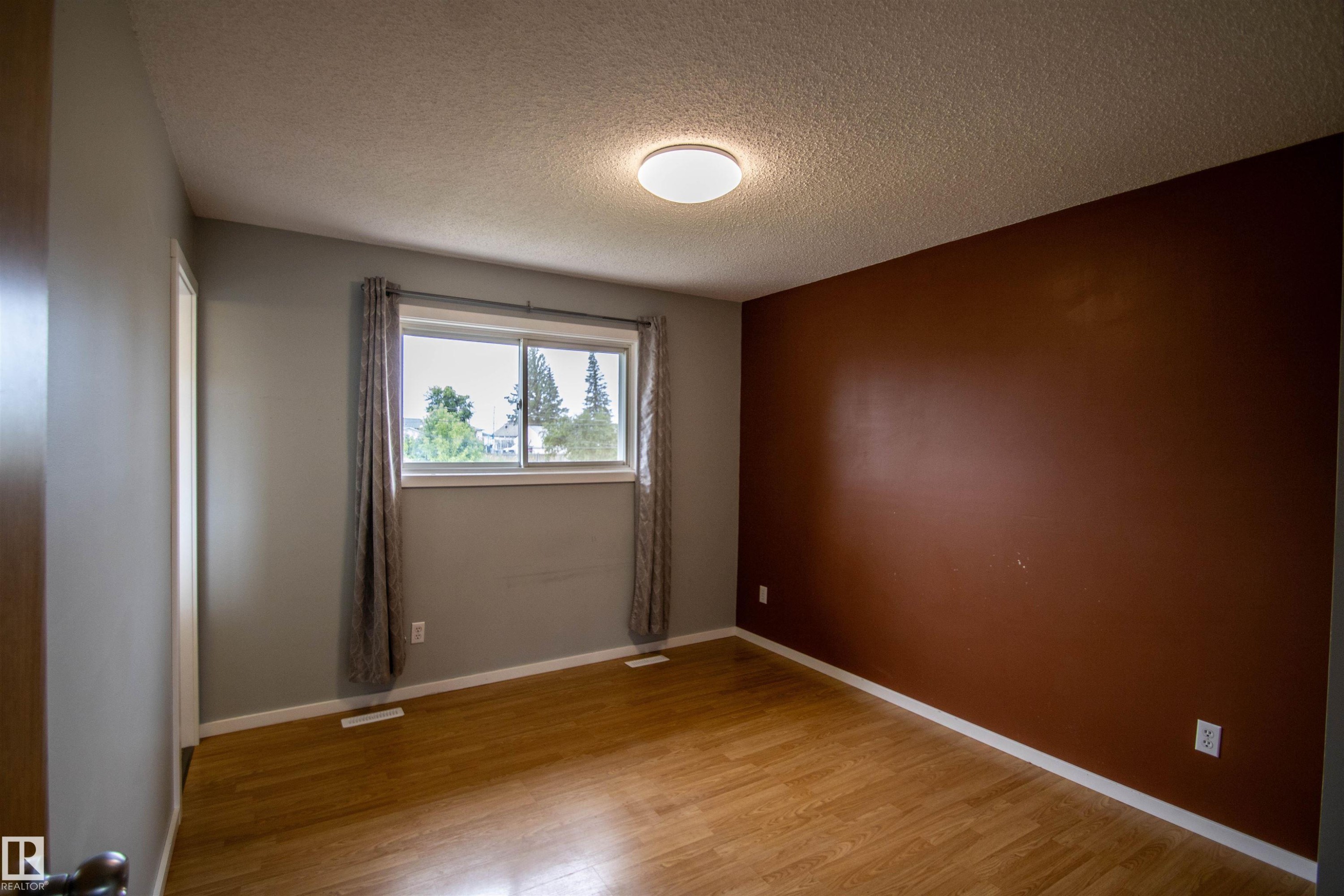 Bright interior space featuring wood-finish flooring, a window with exterior tree line, textured ceiling, and a flush mount ceiling light - 4725 45 Ave, St. Paul Town, AB - Indoor Photo Showing Other Room