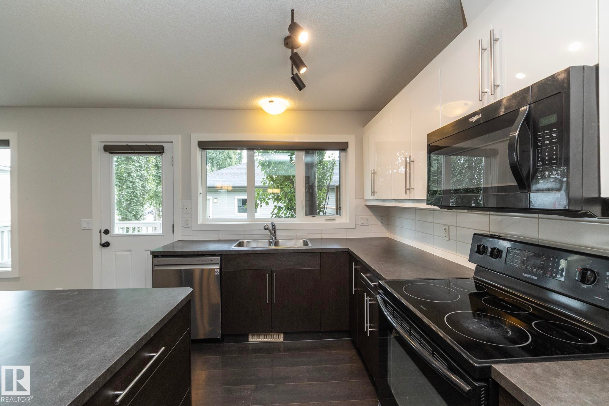 Modern kitchen featuring white upper cabinetry, dark lower cabinetry, stainless steel dishwasher, and black range with microwave - 53/55 Michigan Street, Devon, AB