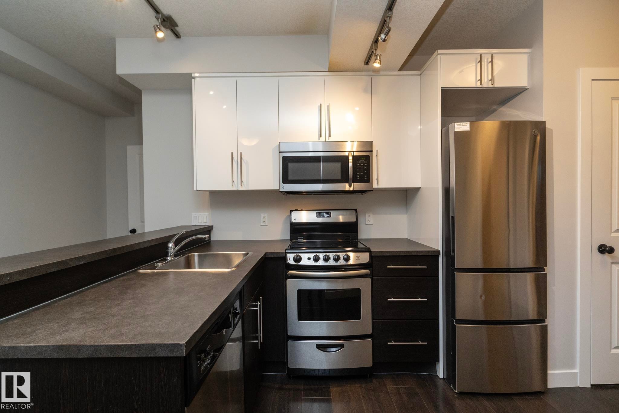 Kitchen featuring dark wood-finish flooring, two-tone cabinetry with white upper cabinets, stainless steel appliances, a dual-level breakfast bar, and track lighting - 53/55 Michigan Street, Devon, AB
