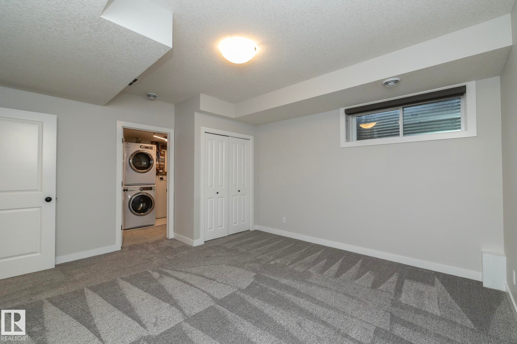 Neutral gray walls with white trim, patterned gray carpet, bi-fold closet, stackable washer and dryer, and a window with blinds - 53/55 Michigan Street, Devon, AB
