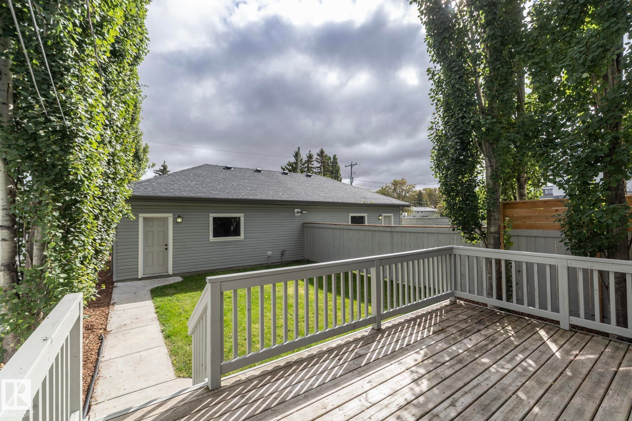 Expansive wood-finish deck with white railings, a concrete pathway, mature trees, and a gray siding structure featuring a dark shingle roof - 53/55 Michigan Street, Devon, AB