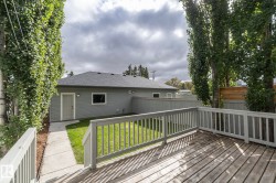 Expansive wood-finish deck with white railings, a concrete pathway, mature trees, and a gray siding structure featuring a dark shingle roof - 