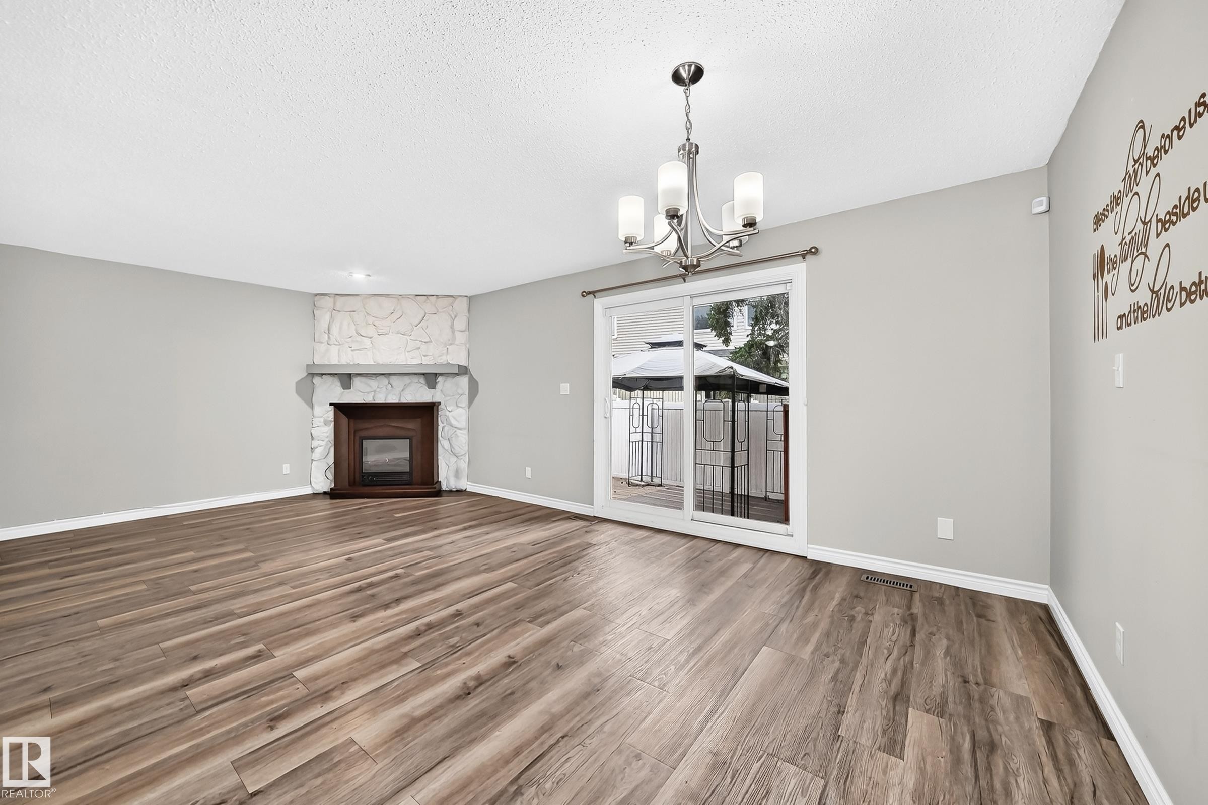 Wood-finish flooring throughout, stone-clad fireplace with wood surround, sliding glass door access to exterior, and a contemporary chandelier - 18146 81 Avenue Nw, Edmonton, AB