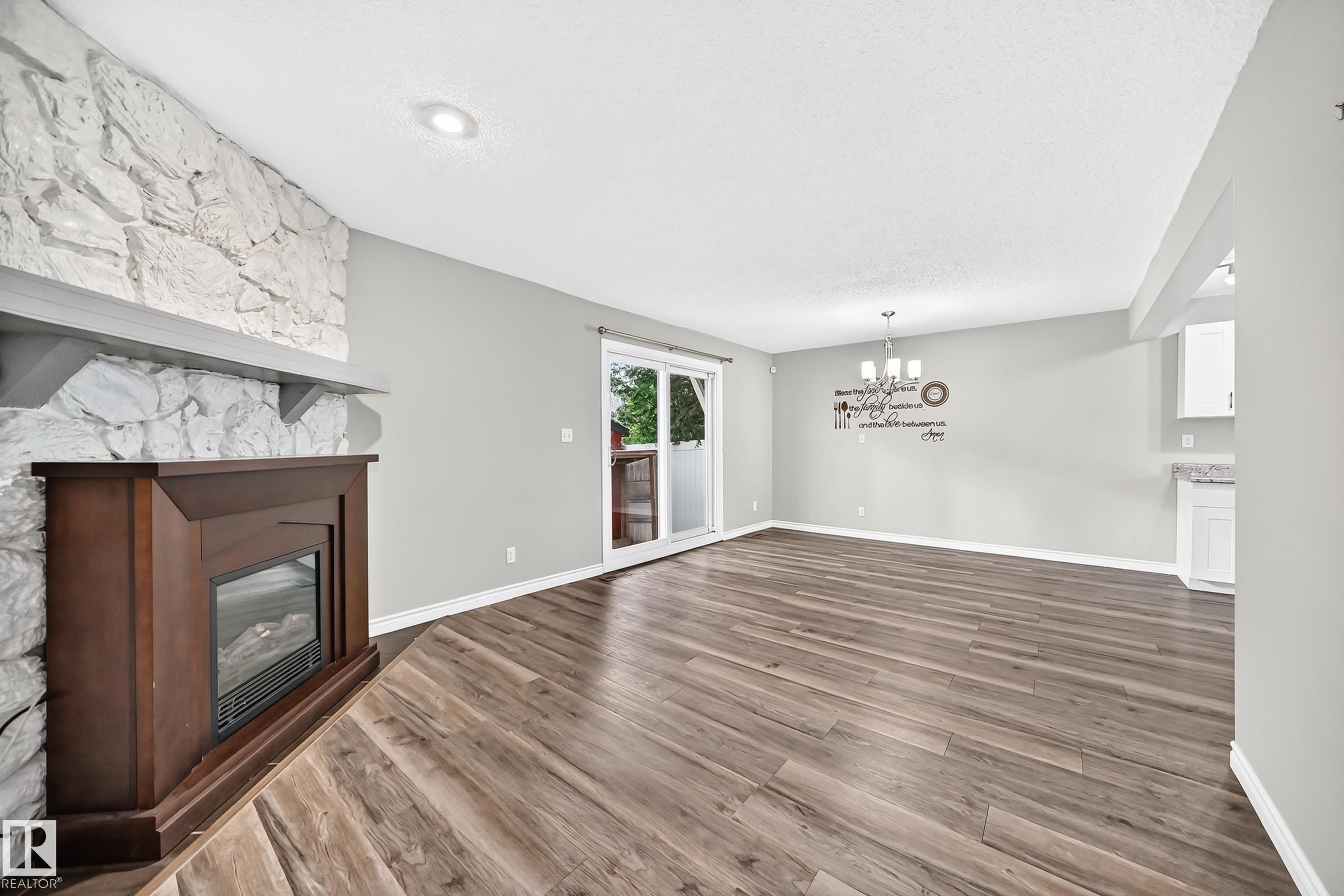 Wood-finish flooring, white stone-clad fireplace surround with wood mantel, built-in electric fireplace, sliding glass door, and a multi-light chandelier - 18146 81 Avenue Nw, Edmonton, AB