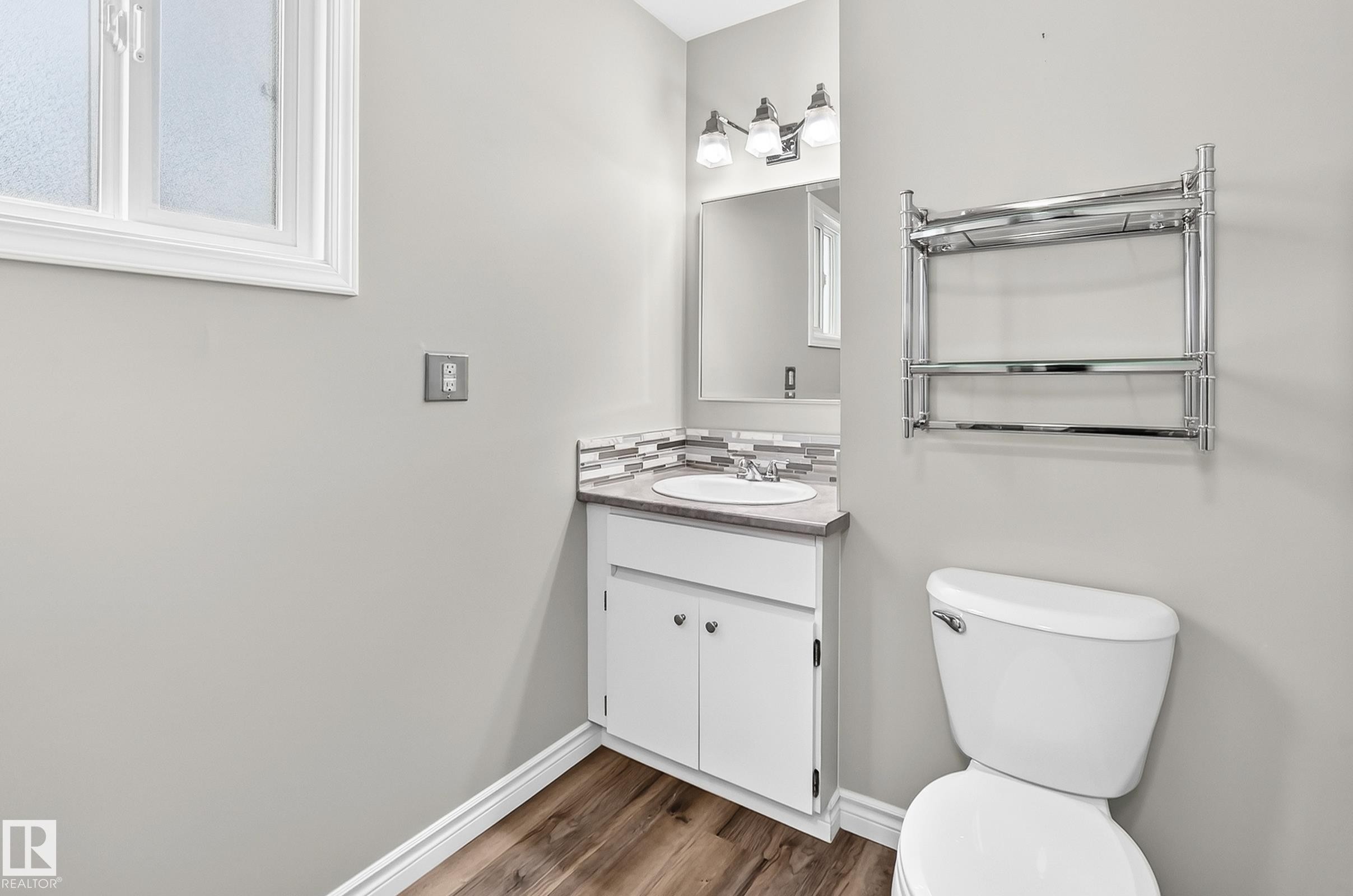 Bathroom with wood-finish flooring, a white vanity with an undermount sink, mosaic tile backsplash, and a three-light vanity fixture - 18146 81 Avenue Nw, Edmonton, AB