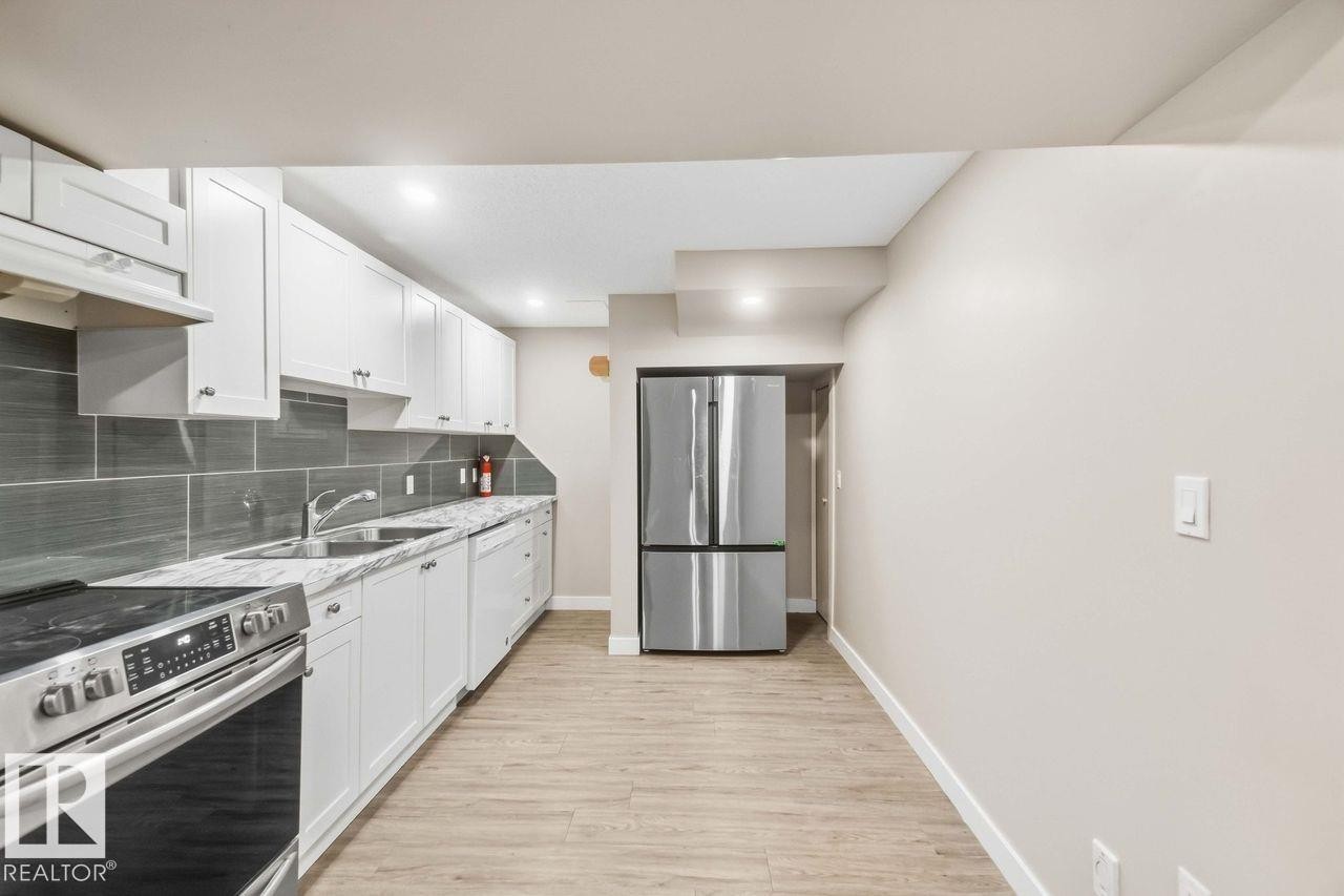 Modern kitchen featuring white cabinetry, gray subway tile backsplash, light-toned wood-finish flooring, stainless steel appliances, and a double-basin sink - 13427 83 Street Nw, Edmonton, AB