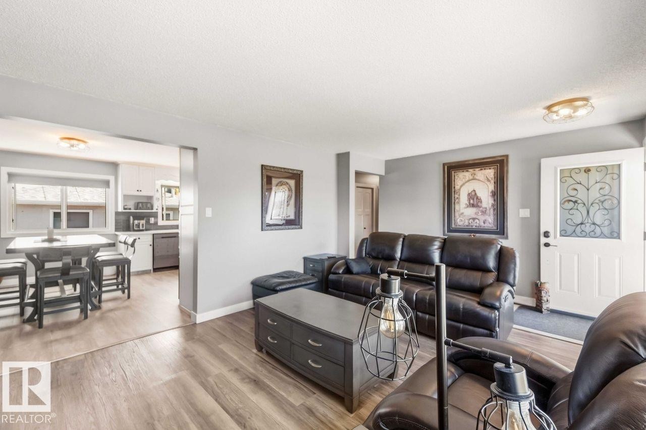 Open-concept living space with wood-finish flooring, a white front door featuring decorative glass, and a recessed kitchen - 13427 83 Street Nw, Edmonton, AB