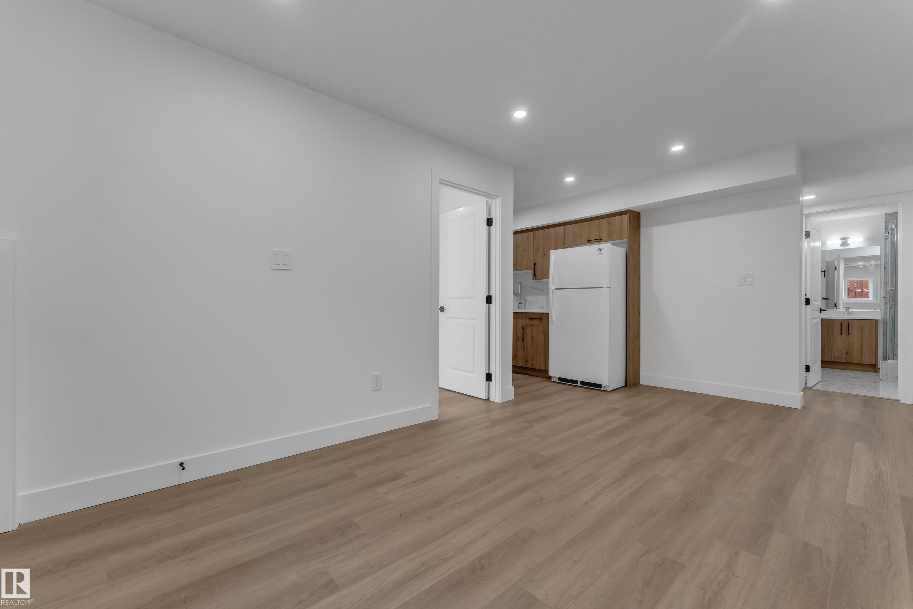 Recessed lighting illuminates wood-finish flooring, white walls, and a kitchen area featuring wood cabinetry, a white refrigerator, and a white tile backsplash - 3320 43 Street Nw, Edmonton, AB