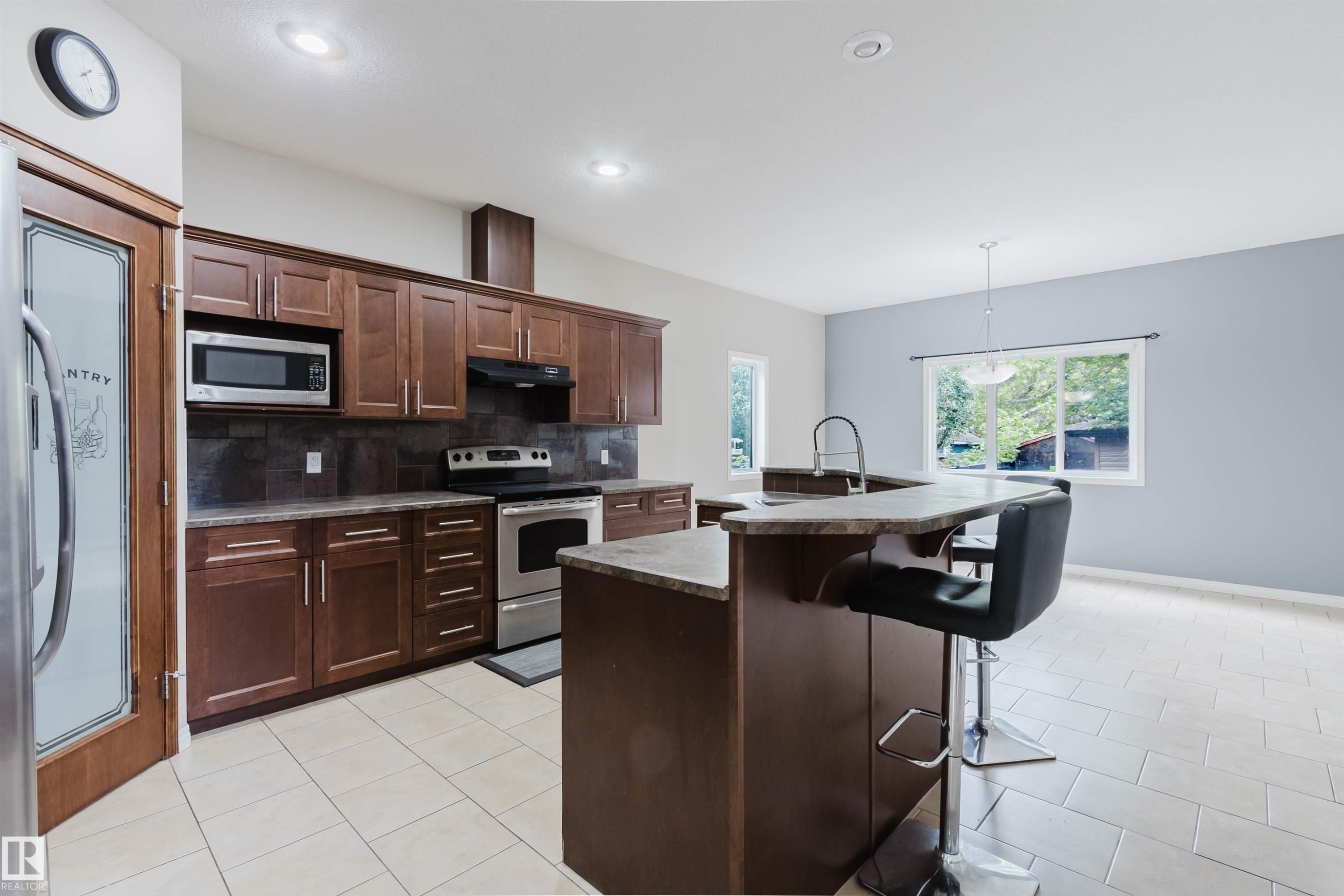 Open-concept kitchen featuring dark wood-finish cabinetry, stainless steel appliances, a stone-finish countertop island with seating, and tile flooring - 11608 96 Street Nw, Edmonton, AB