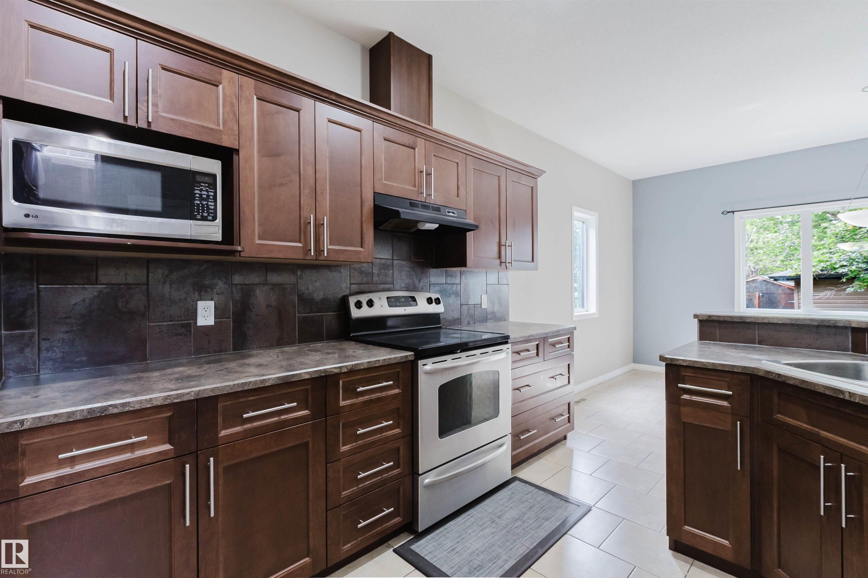 Kitchen featuring rich wood-finish cabinetry, stainless steel appliances, dark tile backsplash, and light tile flooring - 11608 96 Street Nw, Edmonton, AB