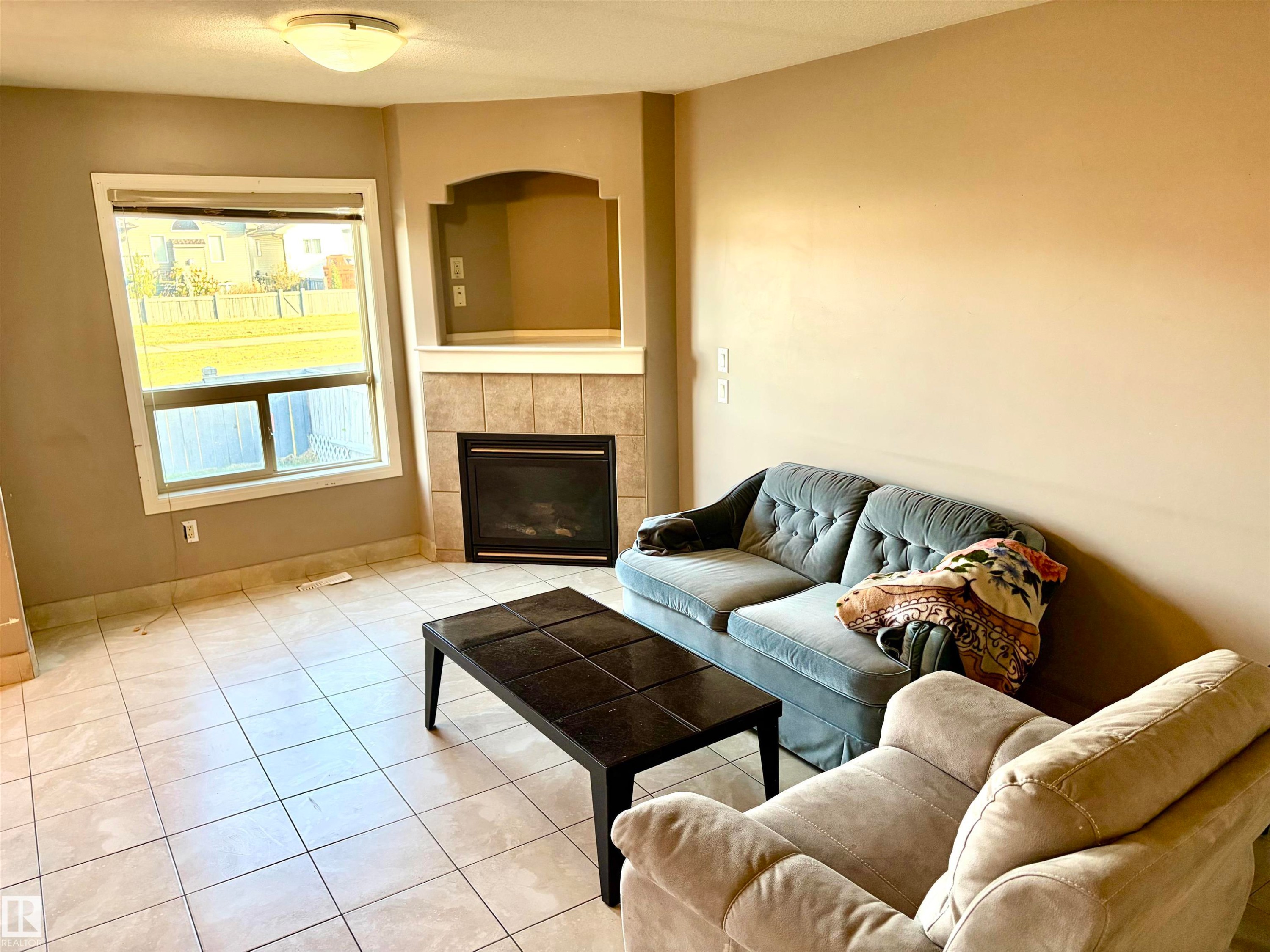 Tiled flooring with a corner fireplace featuring a built-in alcove, large window, and ceiling-mounted light fixture - #17 16317 64 St Nw, Edmonton, AB