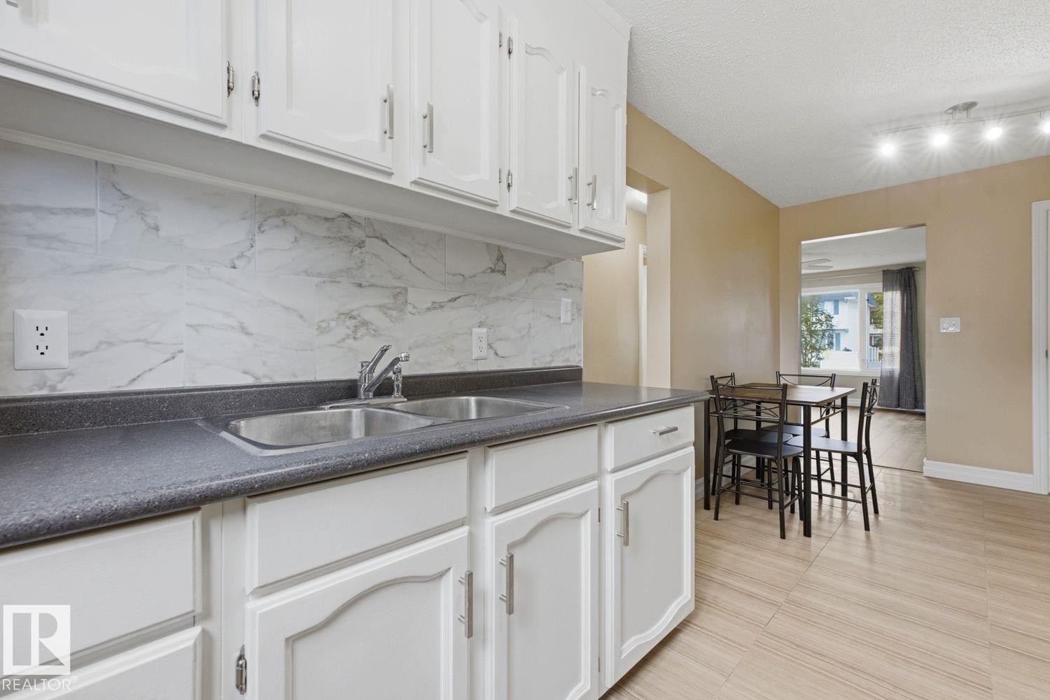 White cabinetry with brushed nickel hardware, a double basin stainless steel sink, a marble-look tile backsplash, and light wood-finish flooring - 11317 22 Avenue Nw, Edmonton, AB