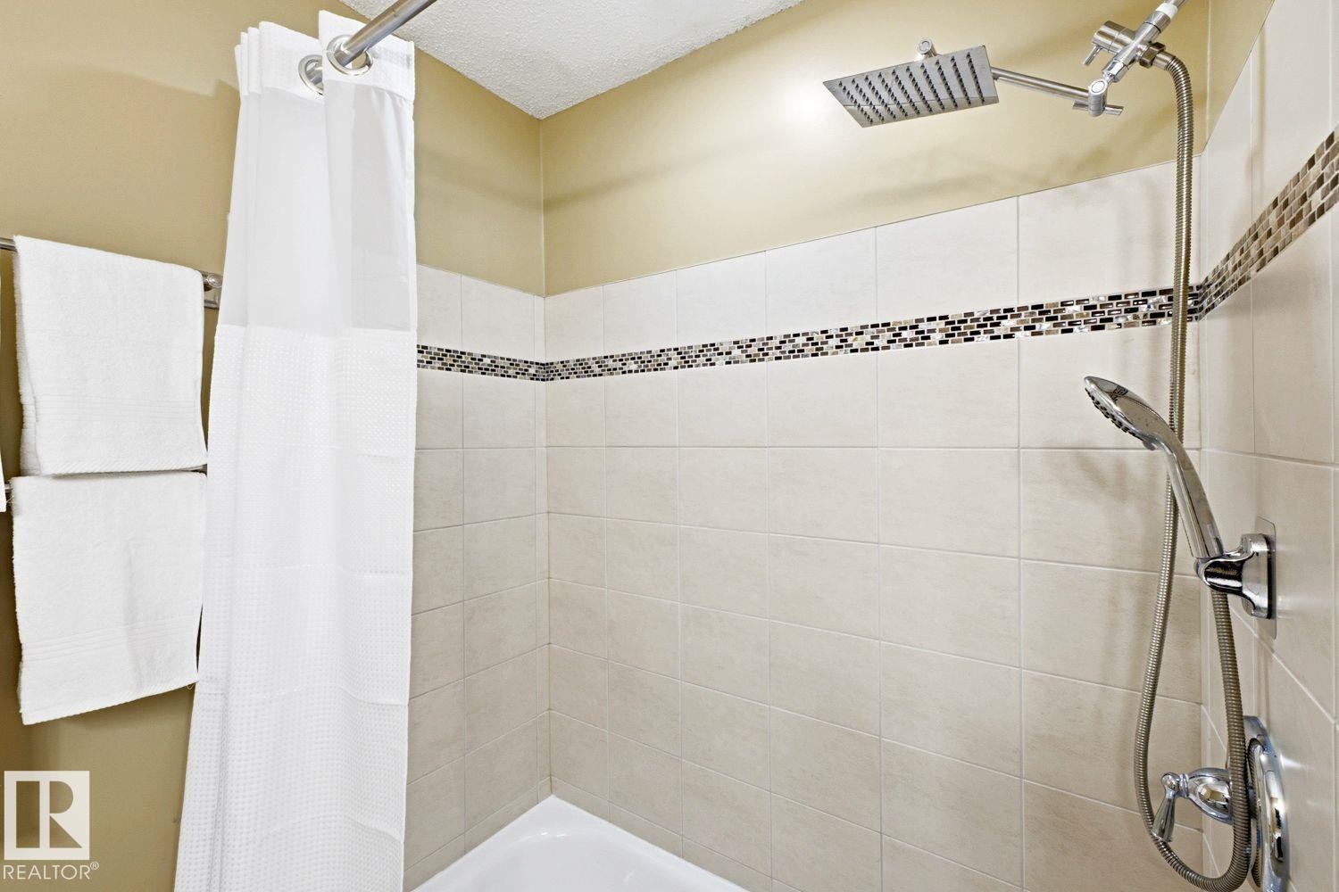Tiled shower enclosure featuring a mosaic accent band, square rain shower head, and an additional handheld shower fixture - 11317 22 Avenue Nw, Edmonton, AB