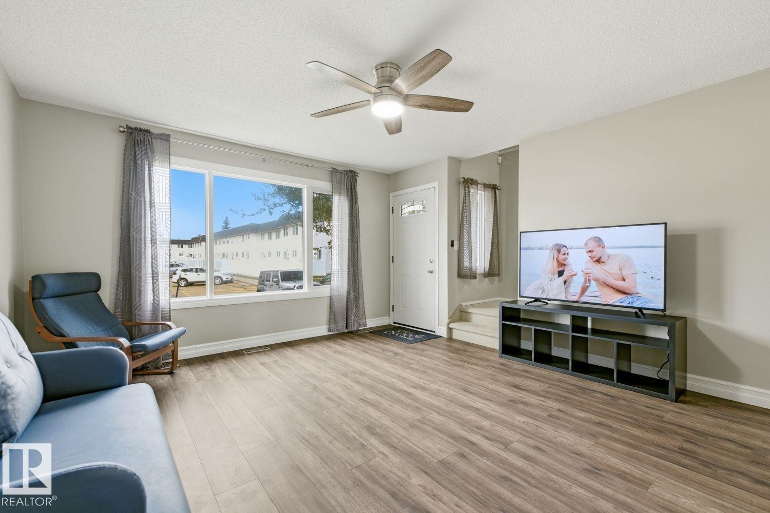 Spacious living area featuring wood-finish flooring, a ceiling fan with light, and a large front window - 11317 22 Avenue Nw, Edmonton, AB