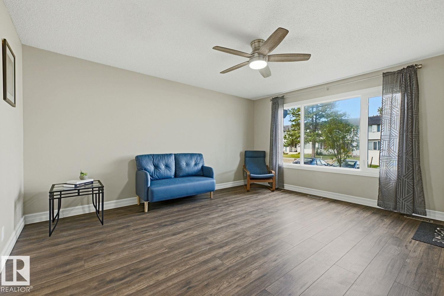 Spacious room featuring wood-finish flooring, a large picture window, and a ceiling fan with integrated lighting - 11317 22 Avenue Nw, Edmonton, AB