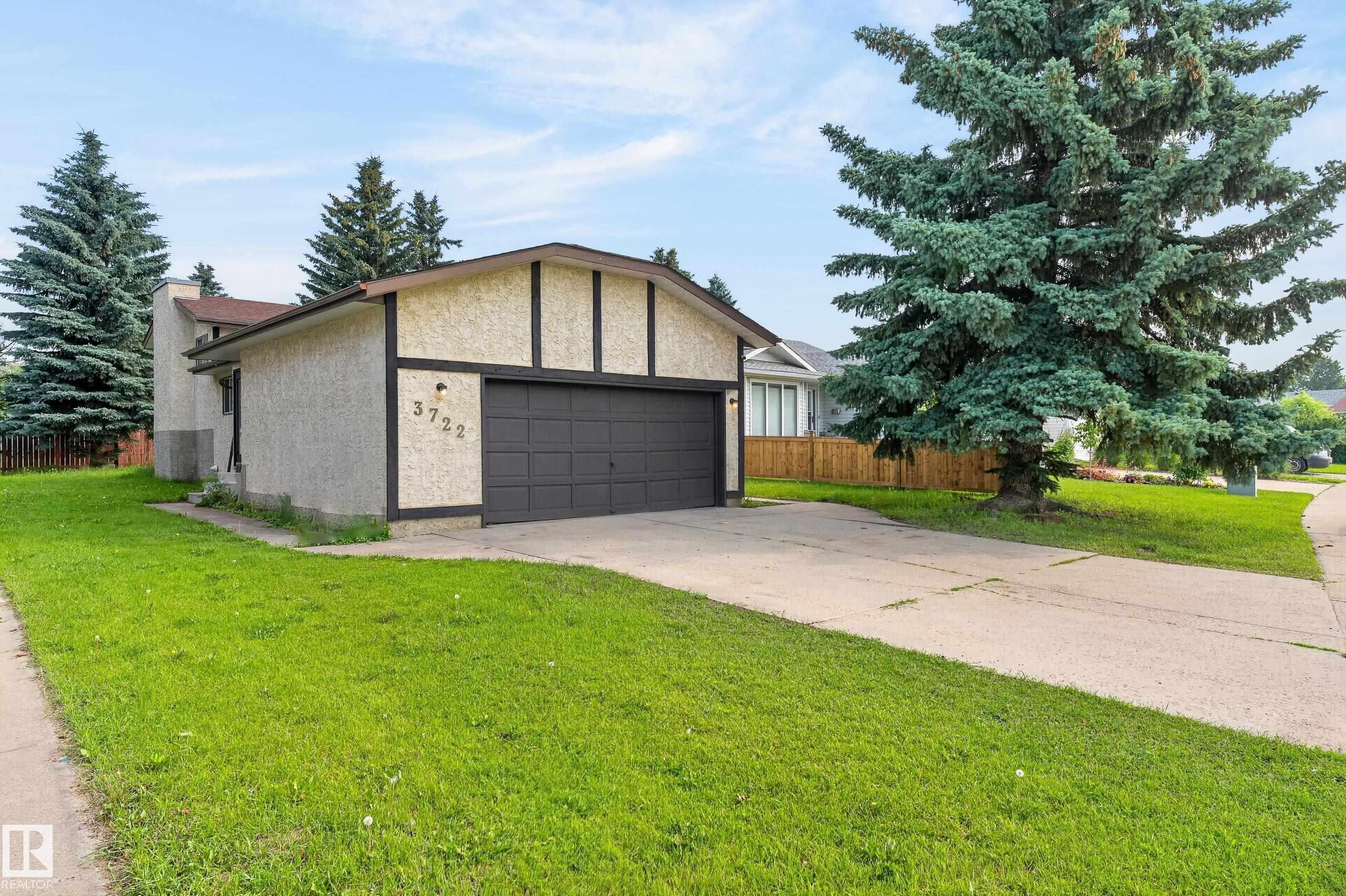 Stucco exterior with contrasting trim, attached garage with dark panel door, concrete driveway, and front lawn - 3722 34A Avenue Nw, Edmonton, AB