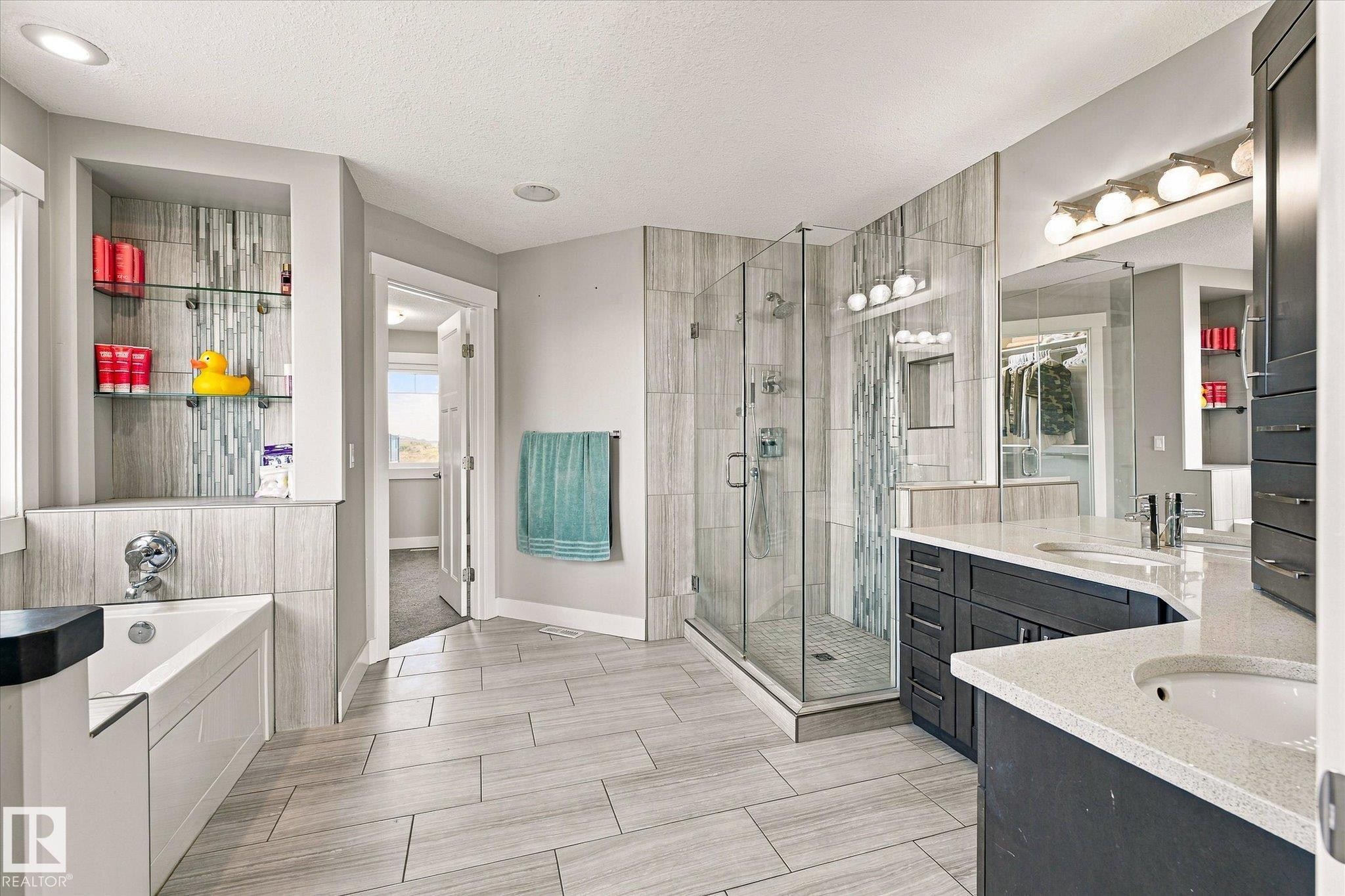 Spacious bathroom featuring wood-finish tile flooring, a walk-in glass shower, dual vanity with undermount sinks, built-in shelving with glass inserts, and a soaking tub - 2011 Genesis Lane, Stony Plain, AB