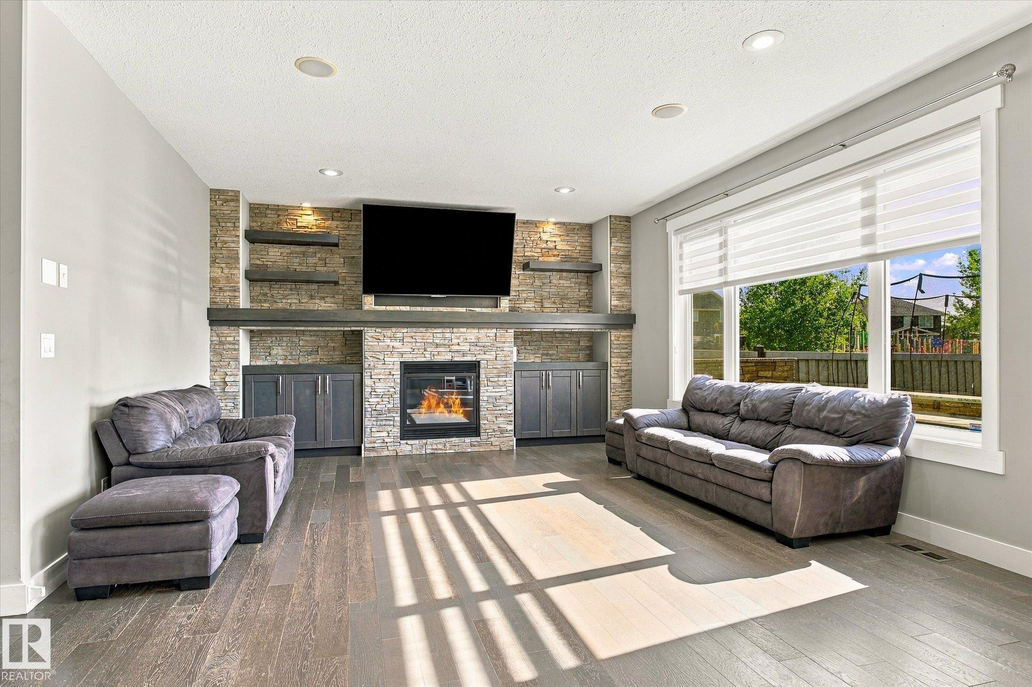 Living area featuring wood-finish flooring, a stone-clad fireplace with built-in cabinetry, floating shelves, recessed lighting, and large windows - 2011 Genesis Lane, Stony Plain, AB