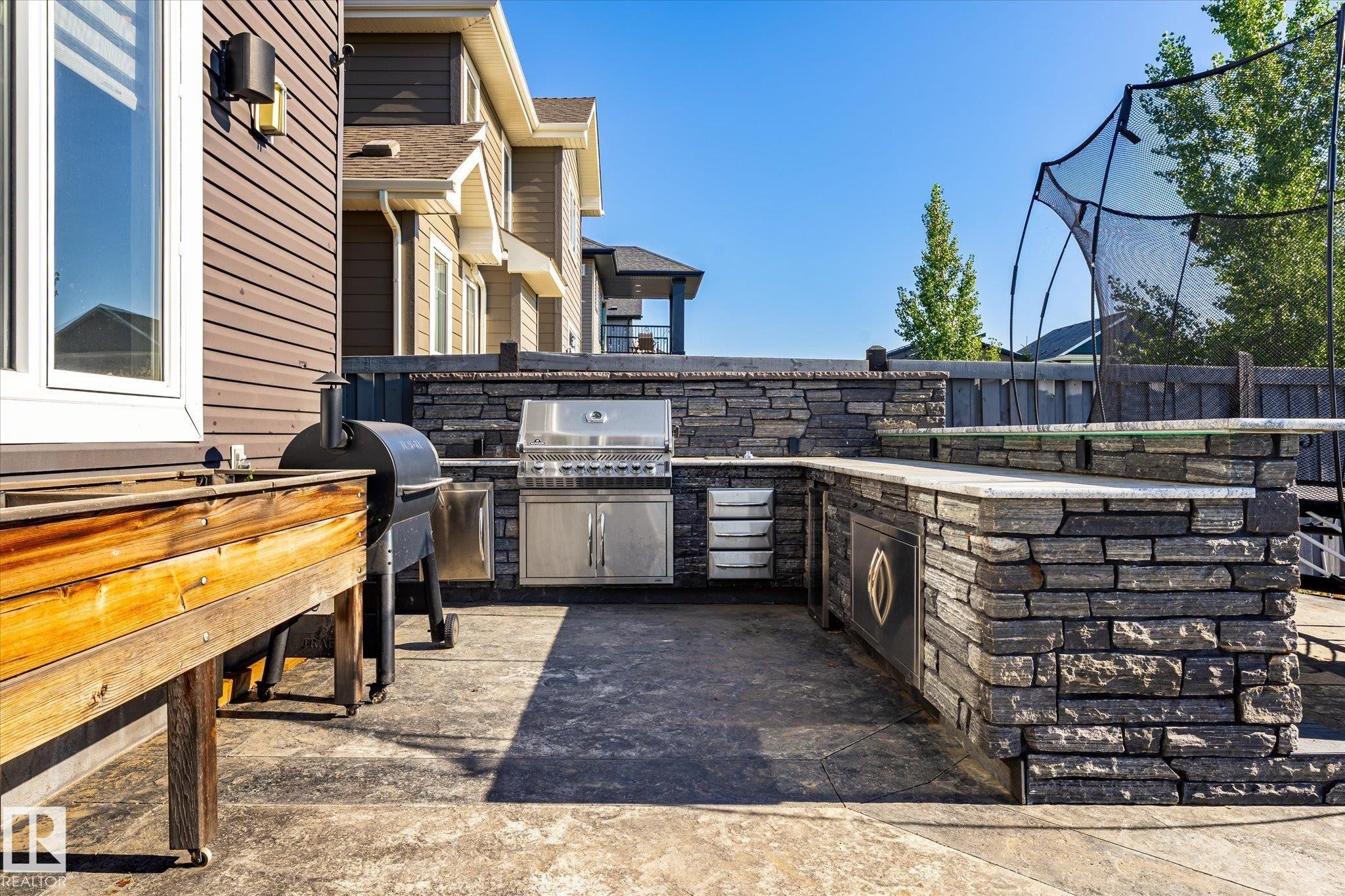 Expansive patio featuring a built-in outdoor kitchen with stainless steel grill, stone veneer base, light-toned countertop, and stamped concrete flooring - 2011 Genesis Lane, Stony Plain, AB