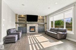 Living area featuring wood-finish flooring, a stone-clad fireplace with built-in cabinetry, floating shelves, recessed lighting, and large windows - 
